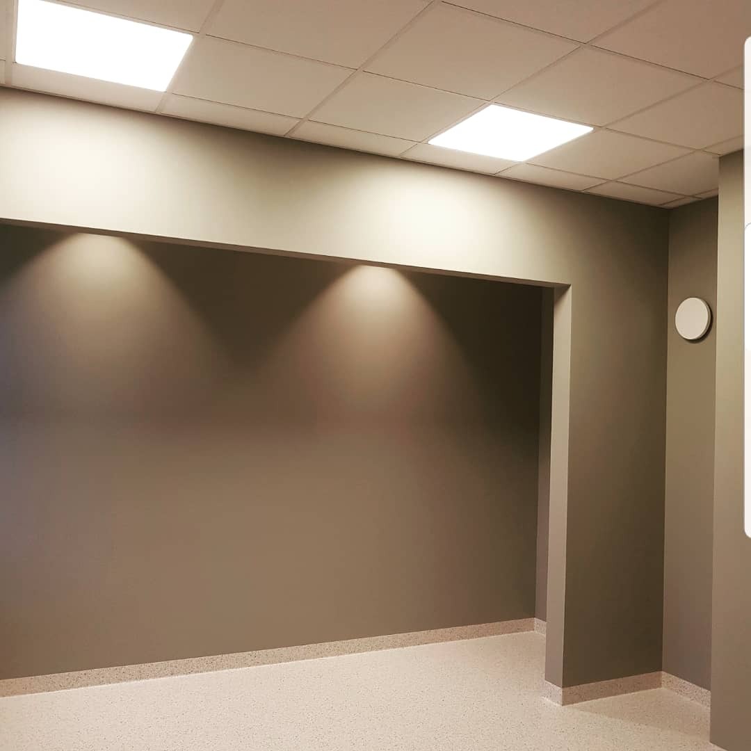 Pomieszczenie biurowe z szarobrązowymi ścianami, oświetleniem punktowym i jasną podłogą z drobnym wzorem, widoczna biała listwa przypodłogowa.