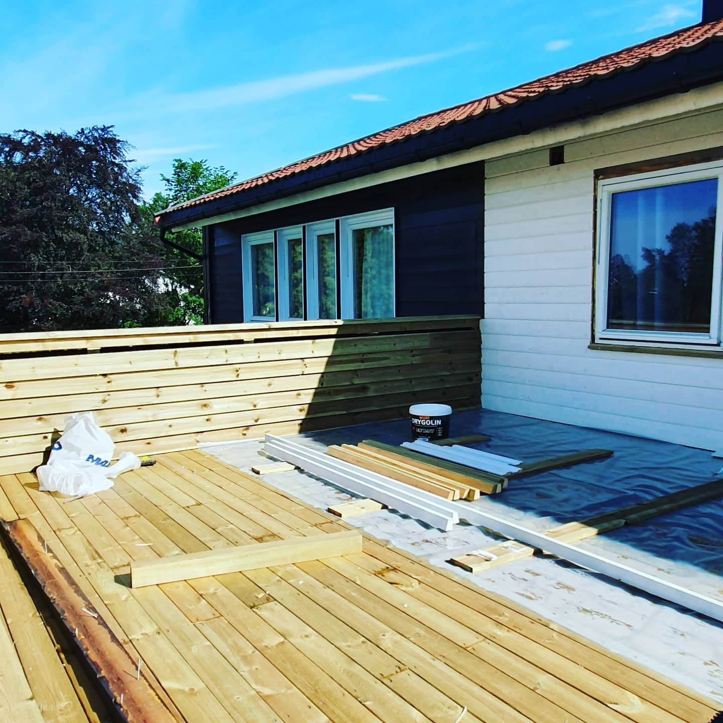 Budowa drewnianego tarasu przy domu z częściowo pomalowaną elewacją; widoczne deski, elementy konstrukcyjne, wiadro z farbą i folia ochronna.