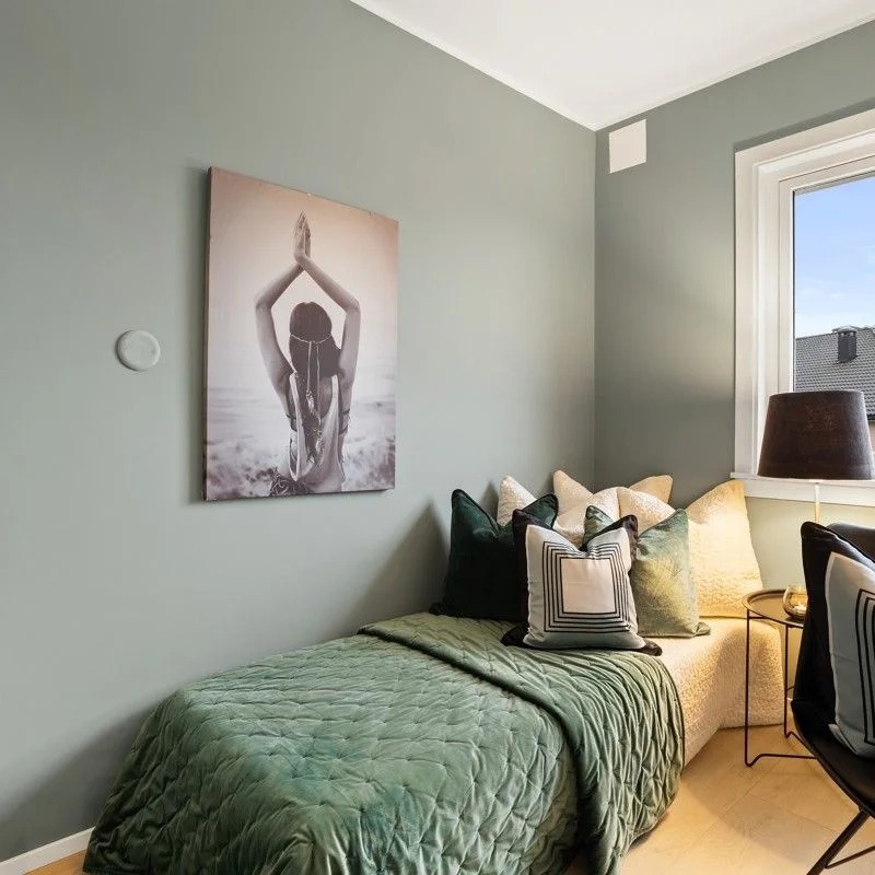 Pokój z łóżkiem jednoosobowym przy ścianie pomalowanej na szaro-zielony odcień, dekorowany obrazem kobiety na plaży i poduszkami w odcieniach zieleni i beżu.
