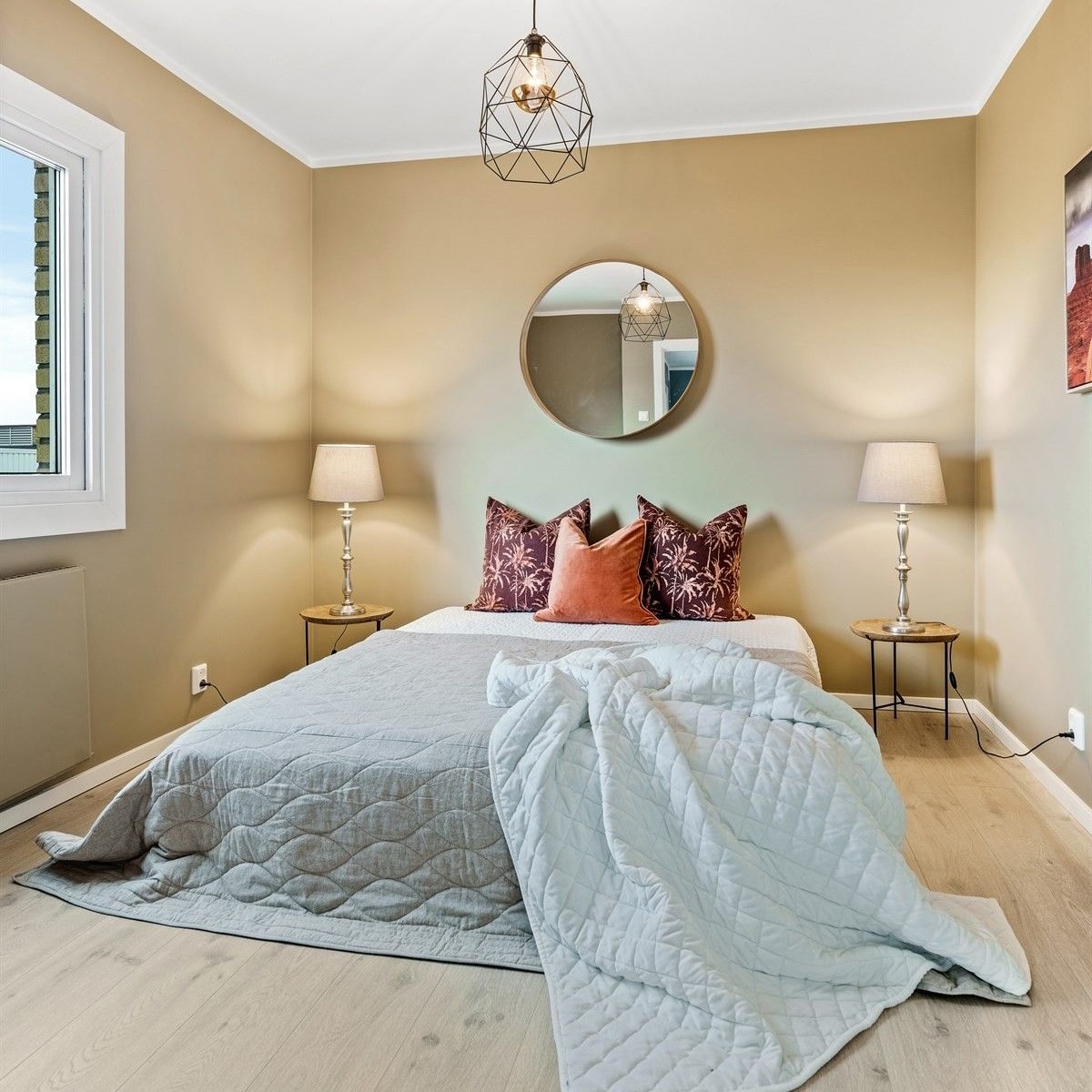 Jasna sypialnia z beżowymi ścianami, drewnianą podłogą, łóżkiem z szarą narzutą i jasnoniebieskim kocem, okrągłym lustrem nad łóżkiem i geometryczną lampą wiszącą.