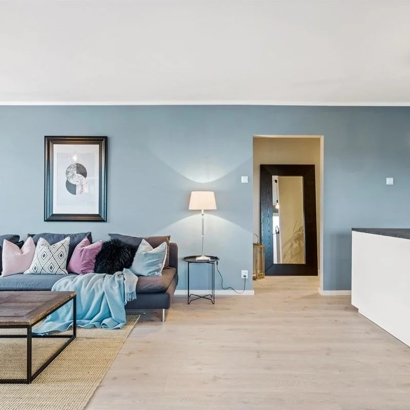 Jasne wnętrze salonu z szarą sofą z poduszkami w odcieniach różu, szarości i czerni, drewnianym stolikiem kawowym, lampą stojącą i dużym lustrem w ciemnej ramie w tle, ściany pomalowane na jasny...