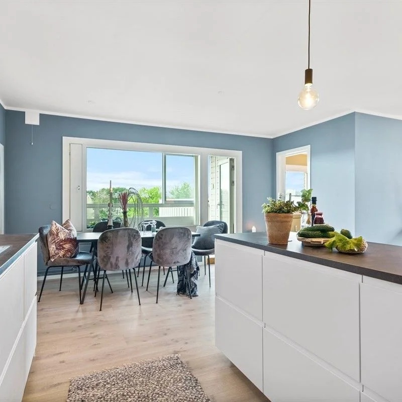 Jasne wnętrze z błękitnymi ścianami, drewnianą podłogą, białymi meblami kuchennymi i stołem jadalnym z aksamitnymi krzesłami, oświetlone wiszącą żarówką edisona.