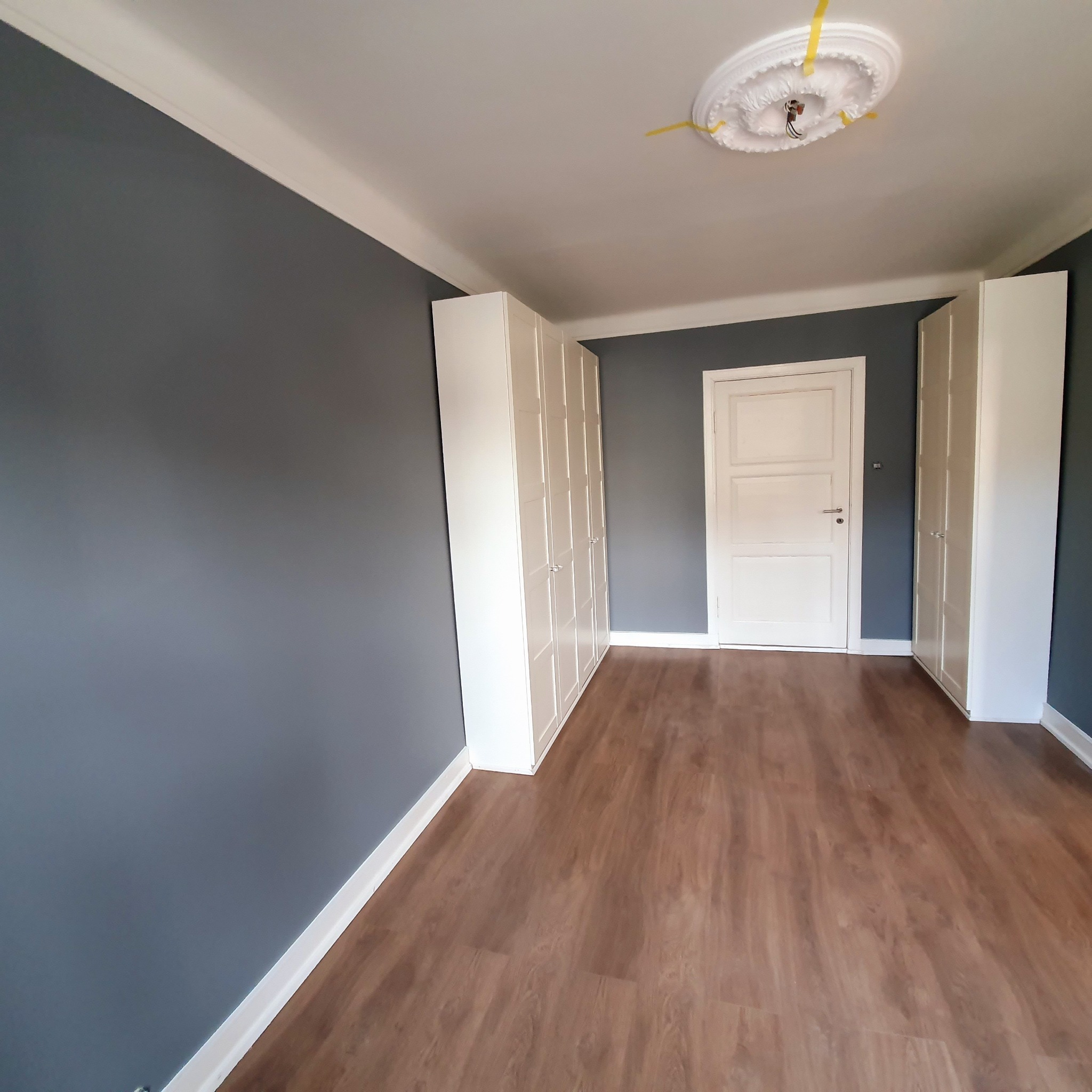 Pokój po remoncie: szare ściany, brązowe panele podłogowe, białe szafy w zabudowie po bokach i białe drzwi na wprost, dekoracyjny element na suficie z wystającymi przewodami.