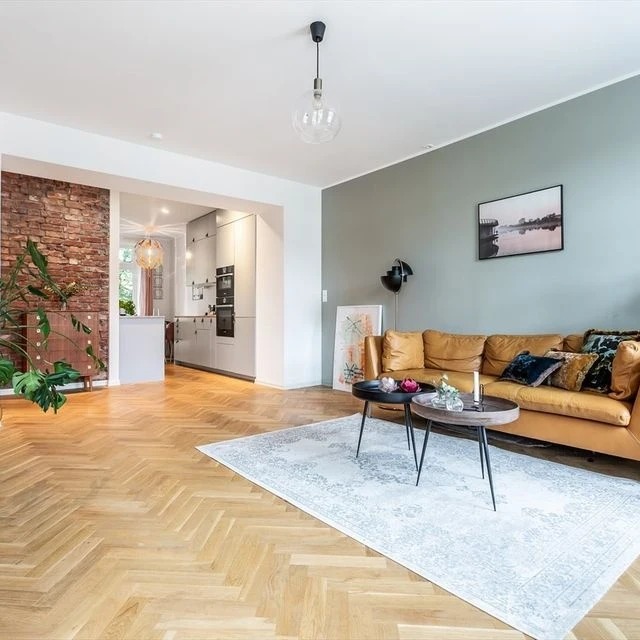 Jasny parkiet ułożony w jodełkę w przestronnym salonie z ceglaną ścianą, skórzaną sofą w odcieniach brązu i widokiem na nowoczesną kuchnię w tle.