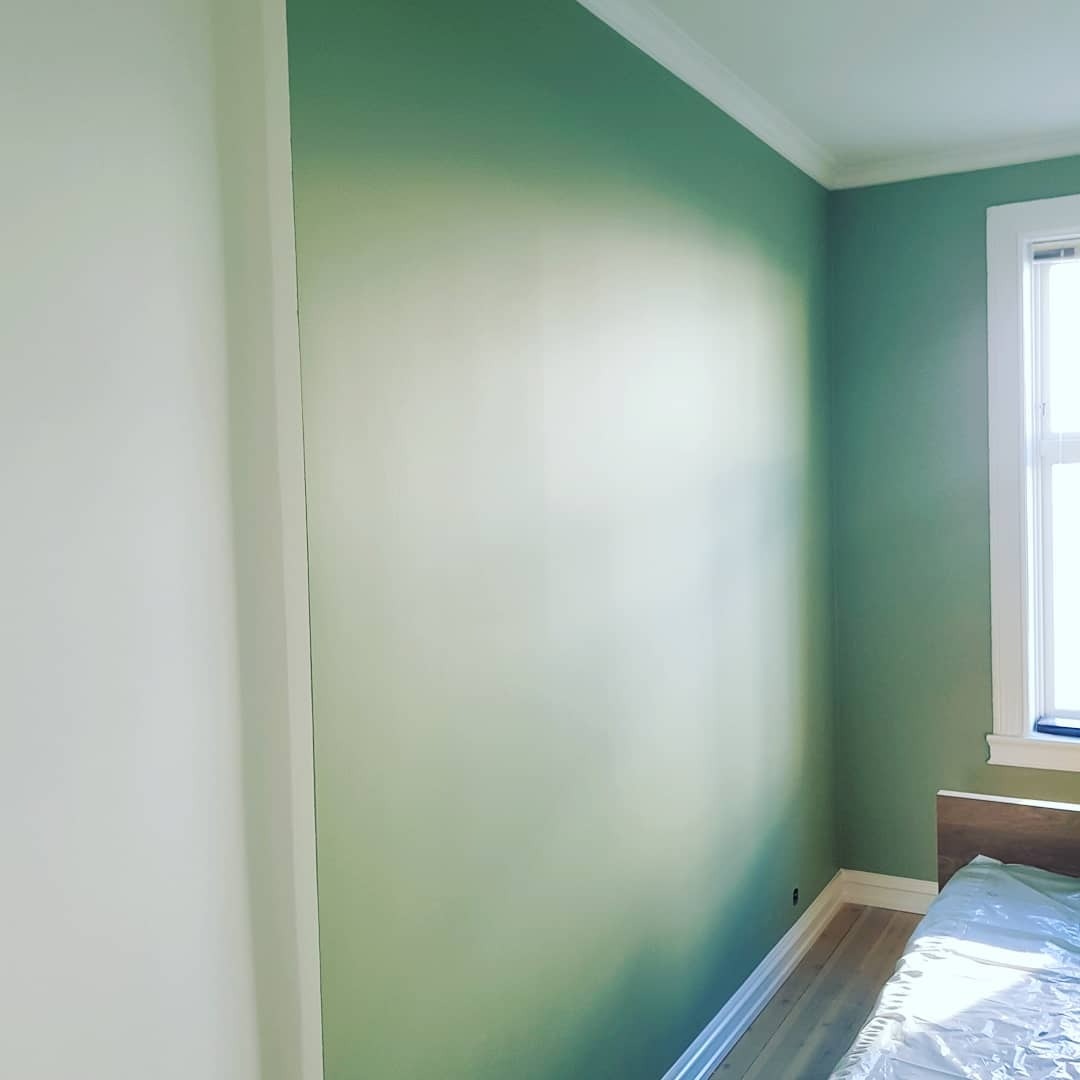 Świeżo pomalowany pokój w odcieniach zieleni, widoczna ściana z delikatnym gradientem koloru, listwy przypodłogowe i okno z białymi ramami, łóżko zabezpieczone folią malarską.
