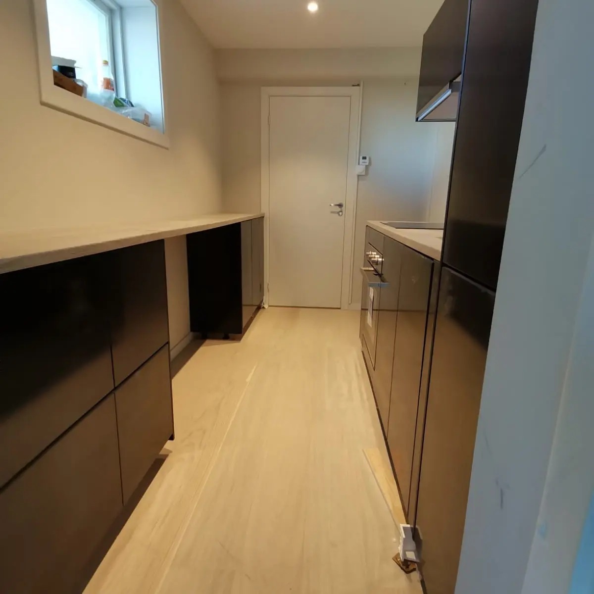 Nowoczesna kuchnia z czarnymi, lakierowanymi szafkami i jasnym, drewnianym parkietem, widok z perspektywy podłogi w kierunku białych drzwi.