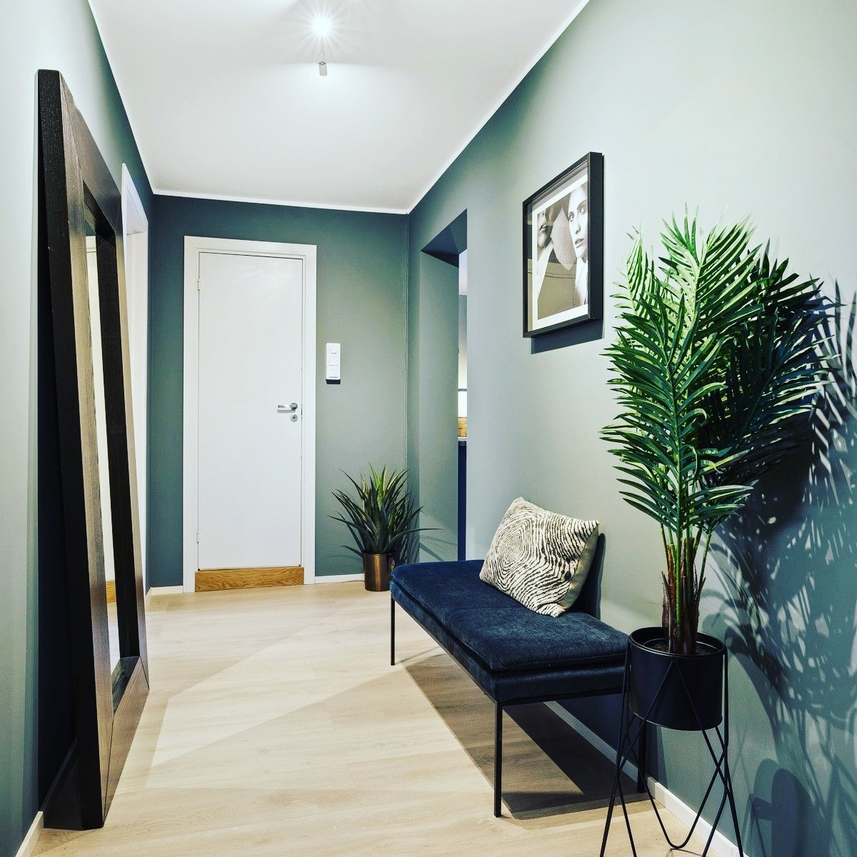 Jasny korytarz z pomalowanymi na szaro ścianami, drewnianą podłogą, ławką z poduszką, lustrem w ciemnej ramie i roślinami w doniczkach.