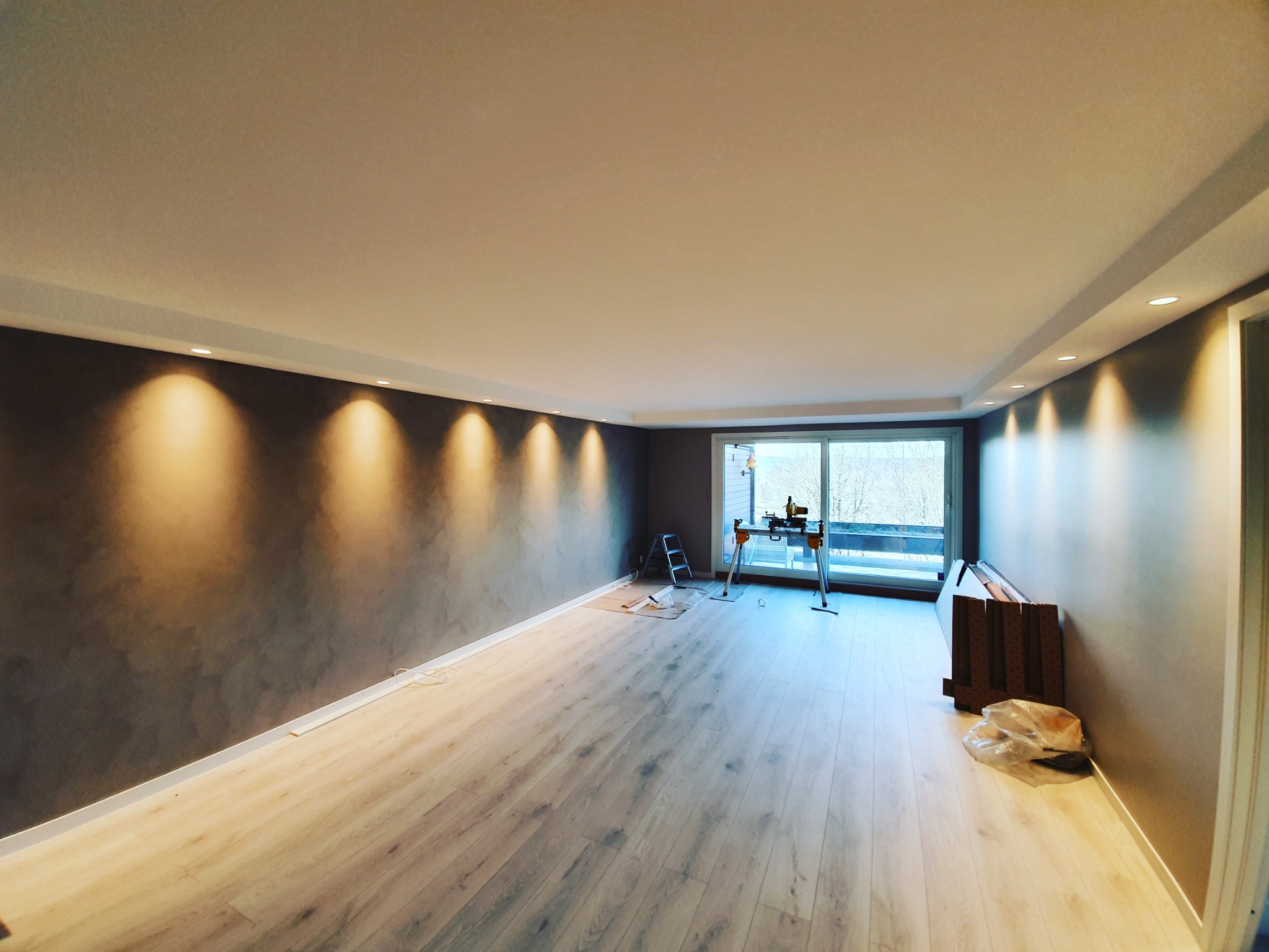 Pokój po remoncie: świeżo ułożona jasna podłoga panelowa, pomalowana na szaro ściana z punktowym oświetleniem, widoczne narzędzia i drabina, przygotowane do dalszych prac wykończeniowych.