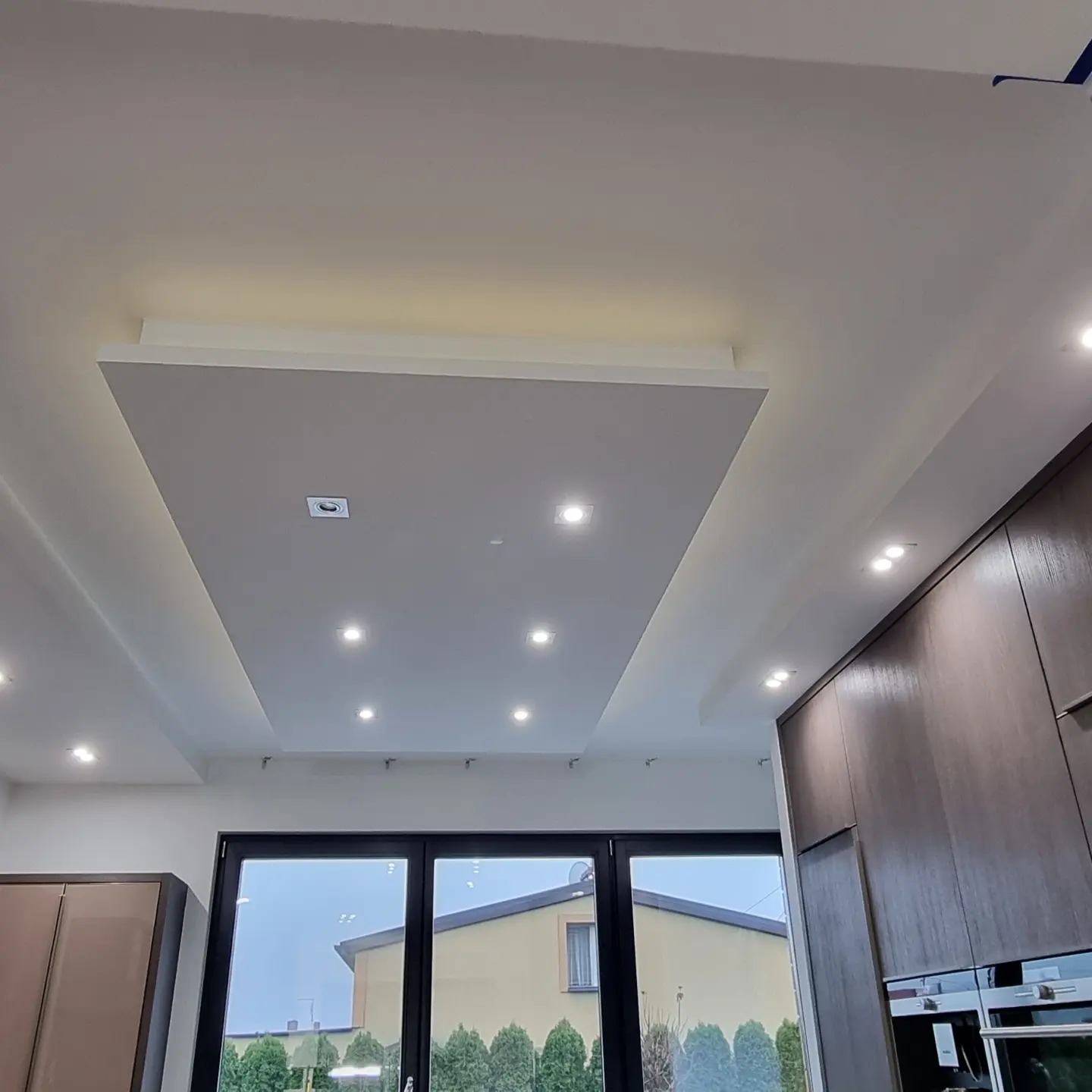 Nowoczesny sufit podwieszany z oświetleniem LED punktowym i listwami LED, widok na okno z zewnętrznym otoczeniem.