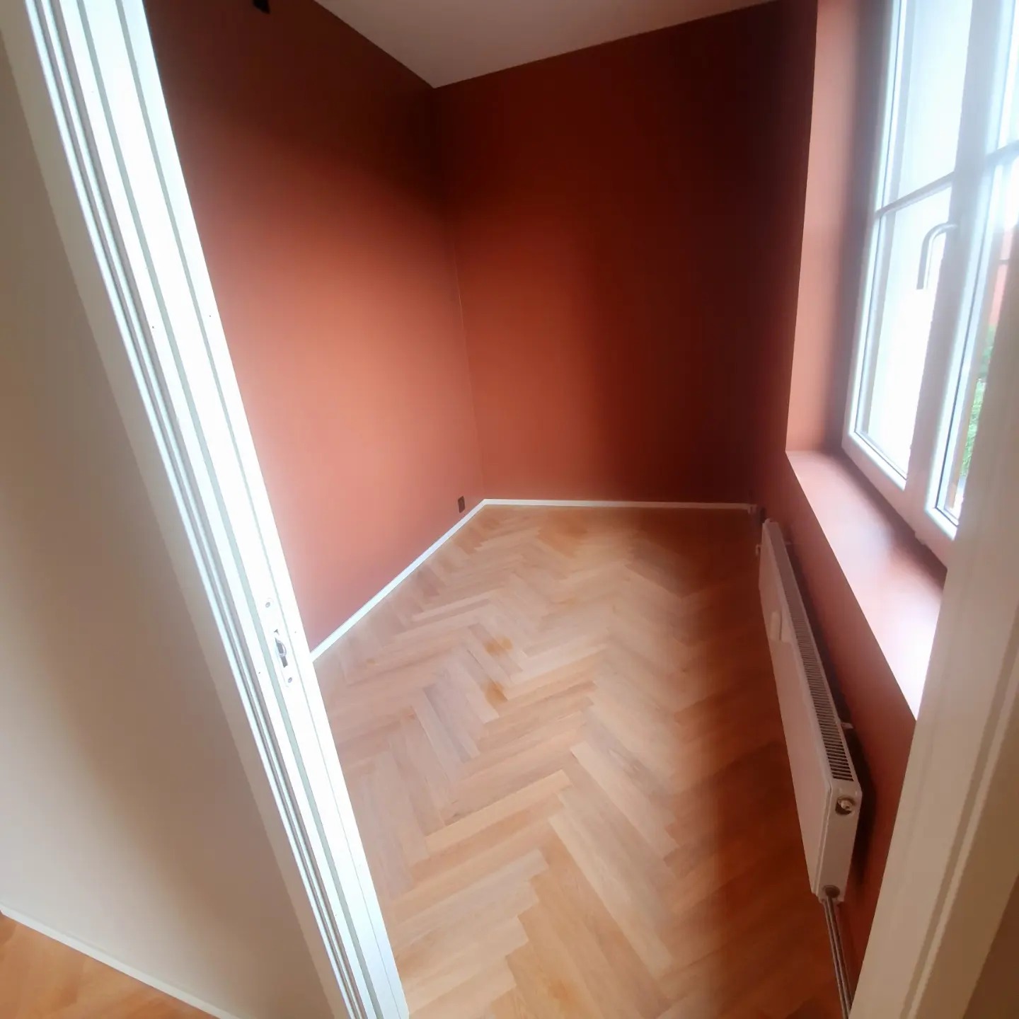 Pomieszczenie po odświeżeniu: ściany w odcieniach terakoty, parkiet ułożony w jodełkę, biały grzejnik pod oknem, widok zza otwartych drzwi.