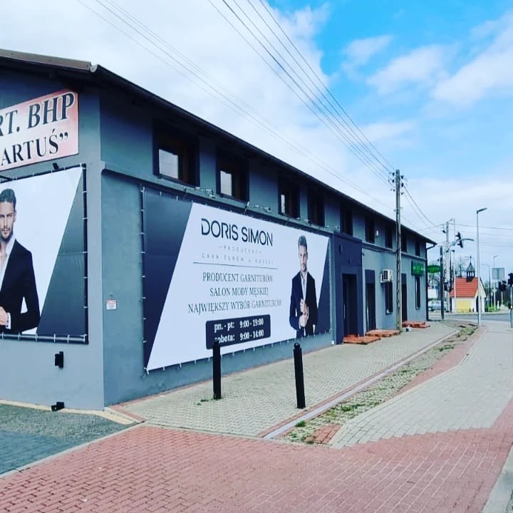Szary budynek z dużymi reklamami salonu mody męskiej Doris Simon, prezentujący producenta garniturów z godzinami otwarcia na banerach.