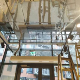 Metalowa ażurowa konstrukcja sufitu podwieszanego z widocznymi elementami instalacji, oświetleniem punktowym i drewnianą drabiną w nowoczesnym biurze z przeszkloną fasadą.