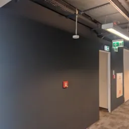 Pomieszczenie biurowe z ciemnoszarą ścianą, widocznymi instalacjami pod sufitem i oświetleniem. Na ścianie zainstalowany czujnik dymu i przycisk alarmowy.