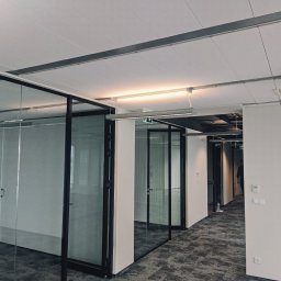 KRAFT-BUDOWNICTWO - Nowoczesne biuro z przeszklonymi ścianami działowymi i panelowym sufitem, widoczne oświetlenie liniowe i szara wykładzina dywanowa.