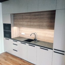 KRAFT-BUDOWNICTWO - Nowoczesna zabudowa kuchenna z szarymi frontami, czarnym blatem i armaturą, oraz drewnopodobnymi szafkami górnymi. Nad blatem widoczne oświetlenie LED.