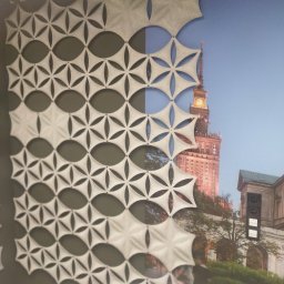 KRAFT-BUDOWNICTWO - Dekoracyjny panel akustyczny o geometrycznym wzorze na ścianie, częściowo zasłaniający fototapetę z widokiem na Pałac Kultury i Nauki w Warszawie.