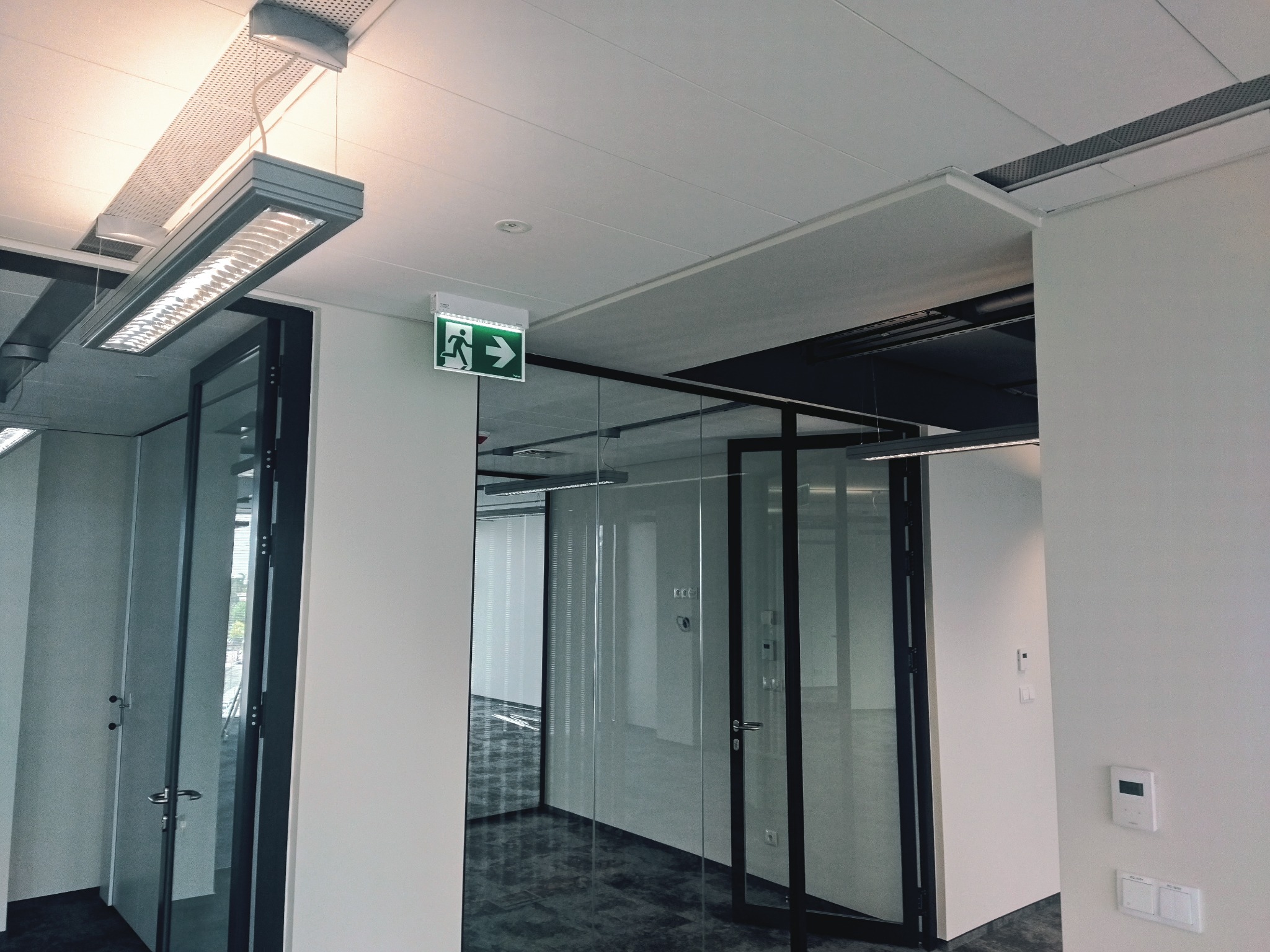 Nowoczesne biuro z przeszklonymi ścianami, widoczne oświetlenie sufitowe i znak ewakuacyjny, minimalistyczny design.