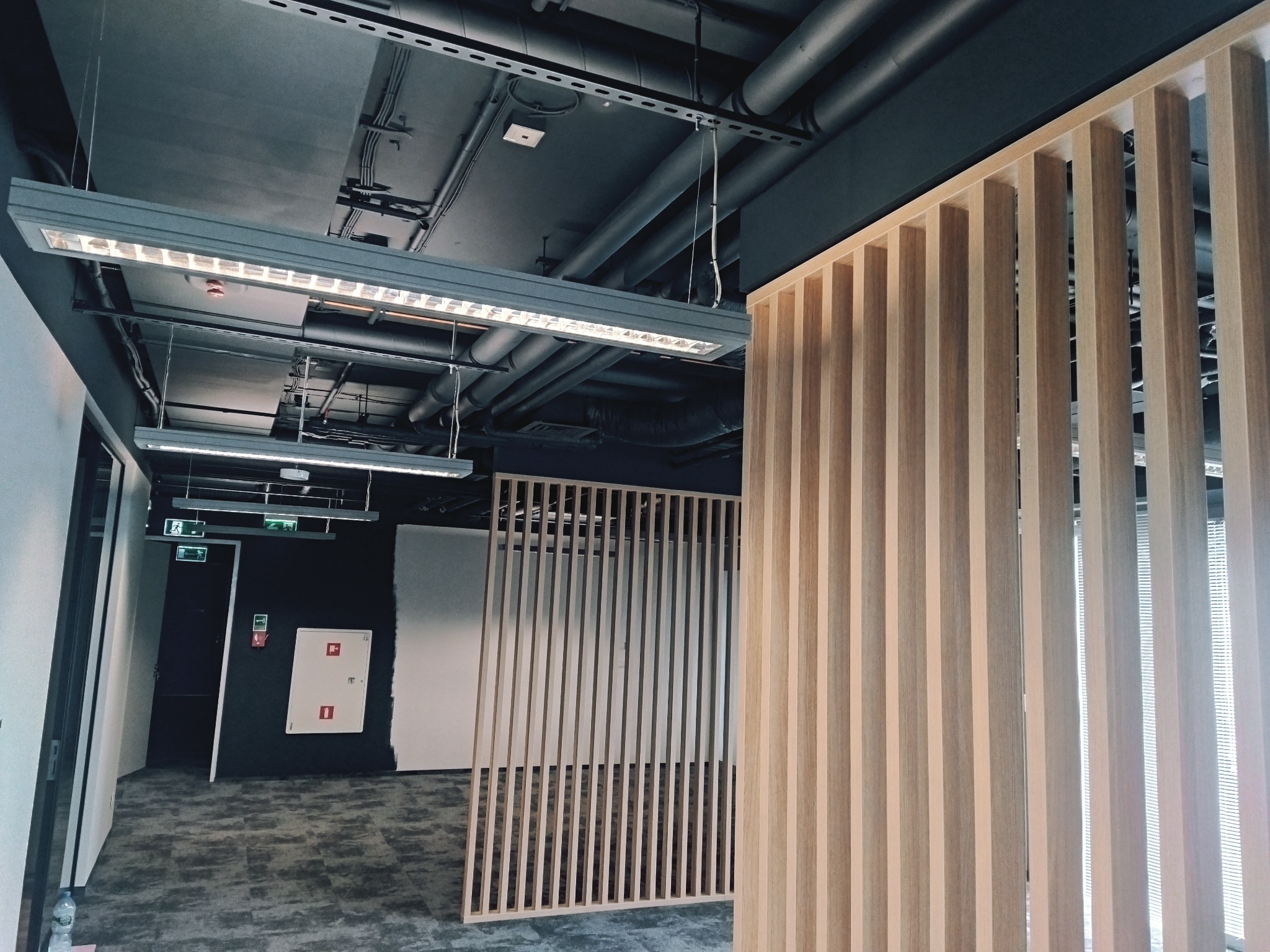 Nowoczesne biuro z drewnianymi lamelami oddzielającymi przestrzeń, widoczne instalacje sufitowe i oświetlenie liniowe.