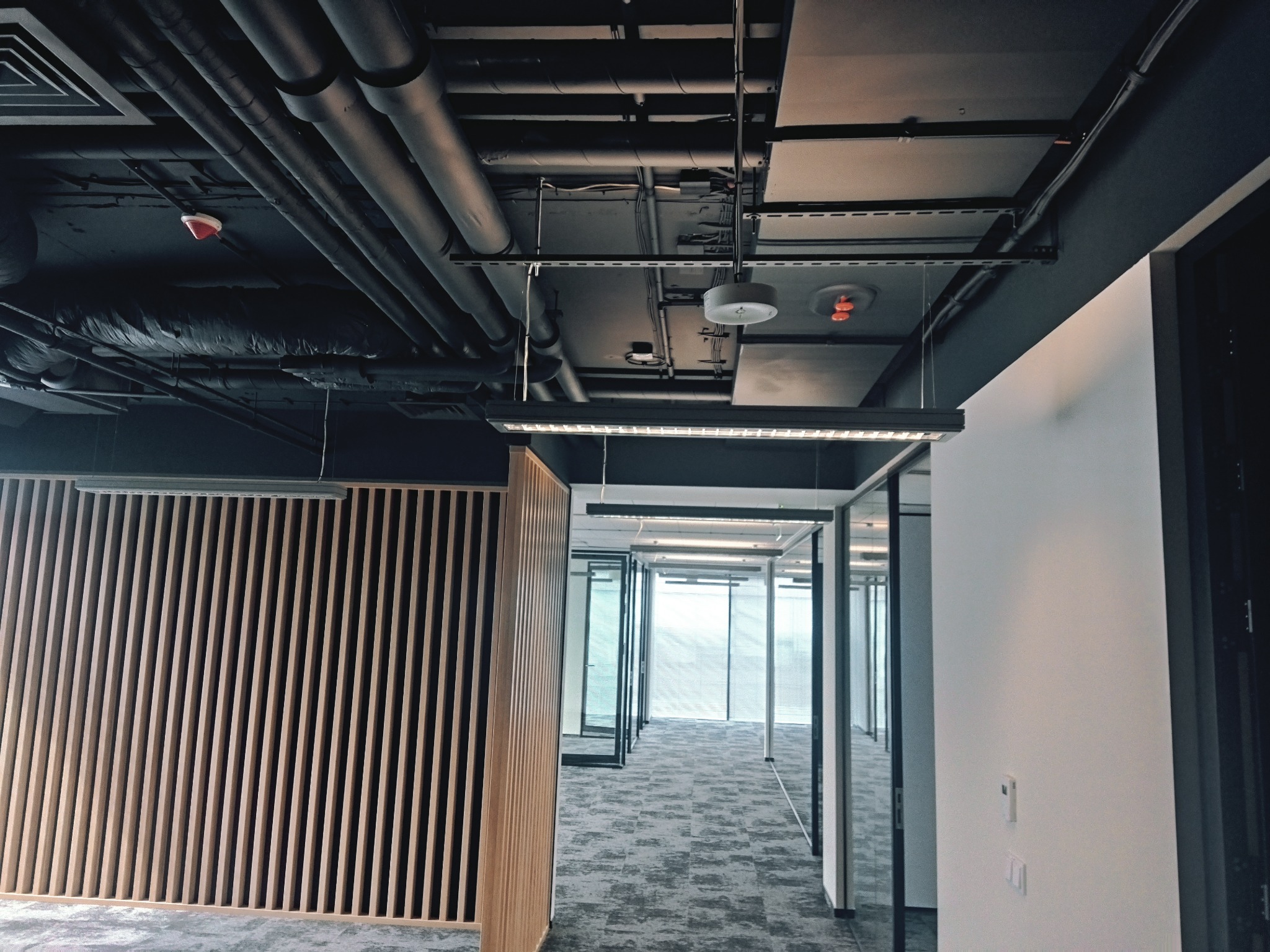 Wnętrze biura po remoncie: widok na korytarz z wykończoną ścianą z drewnianych listew, przeszklone biura z widocznymi żaluzjami, sufit z widocznymi rurami wentylacyjnymi i instalacjami.