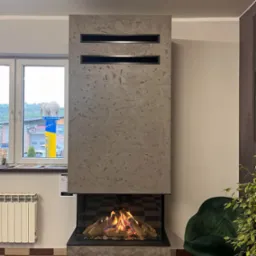Gazowy wkład kominkowy z trójstronnym przeszkleniem zabudowany w płycie krzemianowo-wapniowej wykończonej betonem dekoracyjnym, widoczny płomień i imitacja drewna, na tle okno z widokiem na budynek...