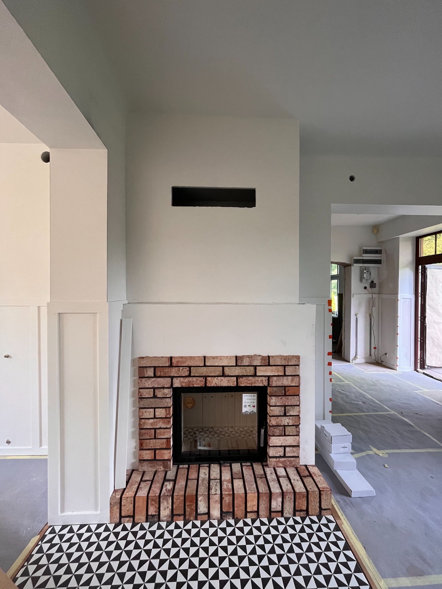 Ceglany portal nowego kominka z czarnym wkładem, obudowany białą ścianą z poziomym otworem wentylacyjnym, podłoga wyłożona czarno-białą mozaiką, widok na wnętrze w trakcie wykończenia.