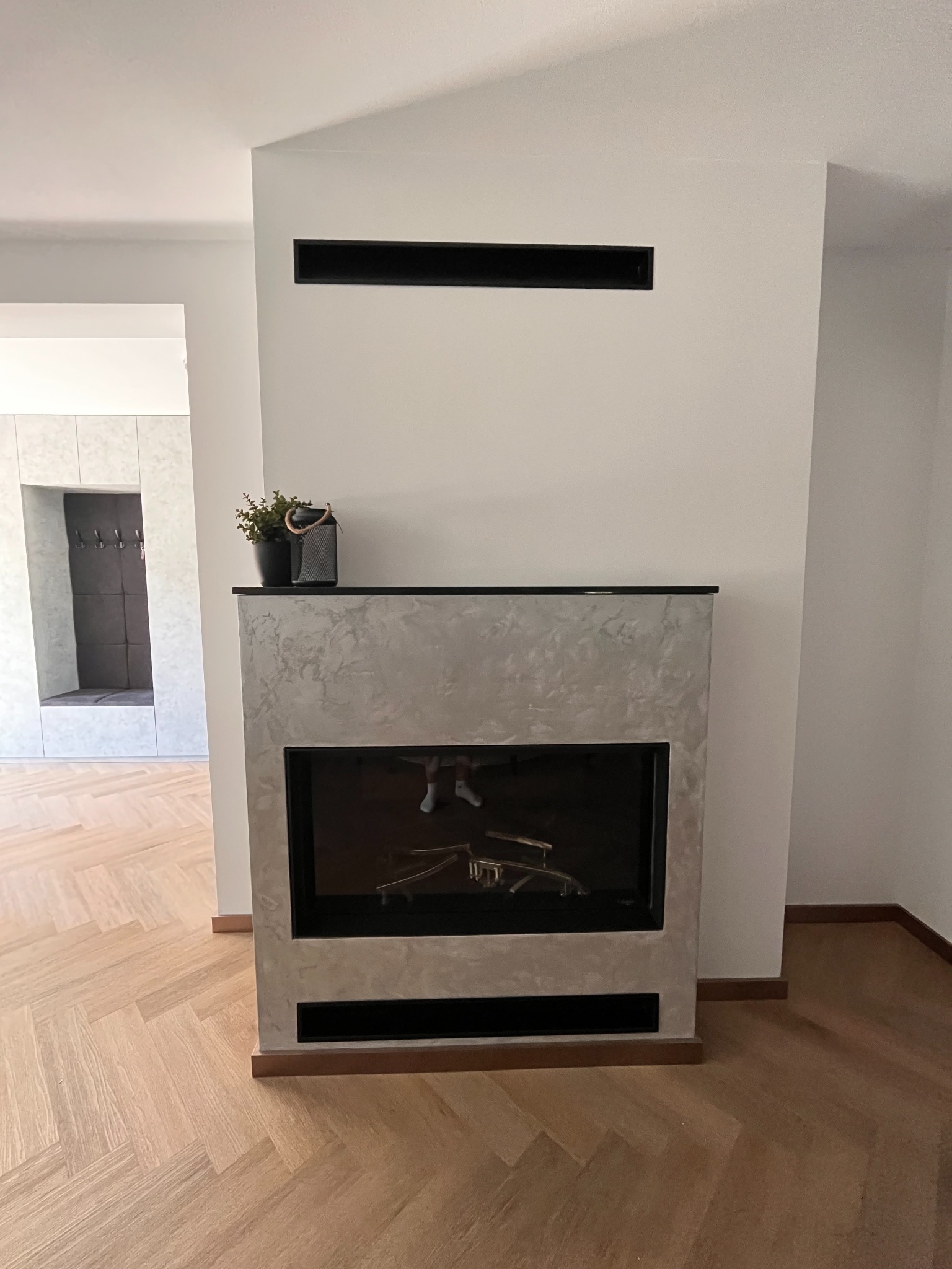 Gazowy kominek Faber Matrix 1050/500 I HPL z obudową w surowym wykończeniu, bez polan, z widoczną kratką wentylacyjną nad nim i parkietem w jodełkę na podłodze.