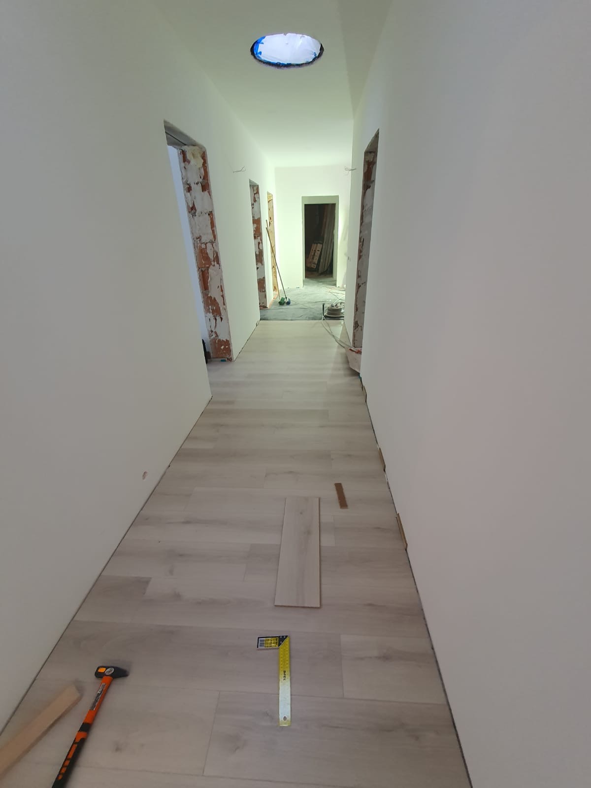 Korytarz w trakcie wykańczania: ułożona podłoga z paneli, narzędzia pomiarowe i młotek leżą na podłodze, widoczne niedokończone ościeżnice drzwiowe.