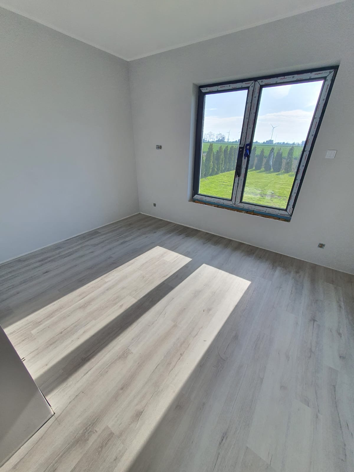 Jasne panele podłogowe ułożone w pustym pokoju z oknem wychodzącym na zielone pole z drzewami i turbiną wiatrową w oddali, oświetlone promieniami słońca.
