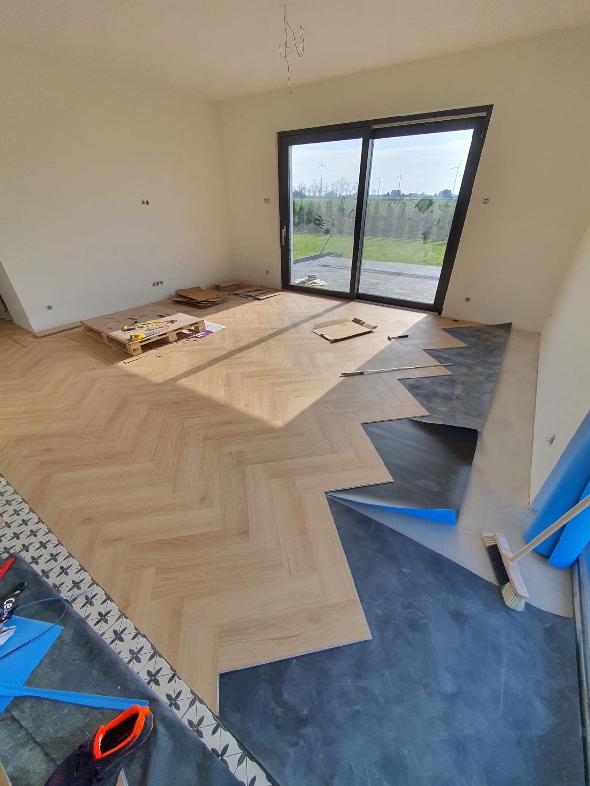 Układanie jasnych paneli podłogowych w jodełkę w nowym domu, widoczne narzędzia na palecie, podkład pod panele i zamiatacz do kurzu.