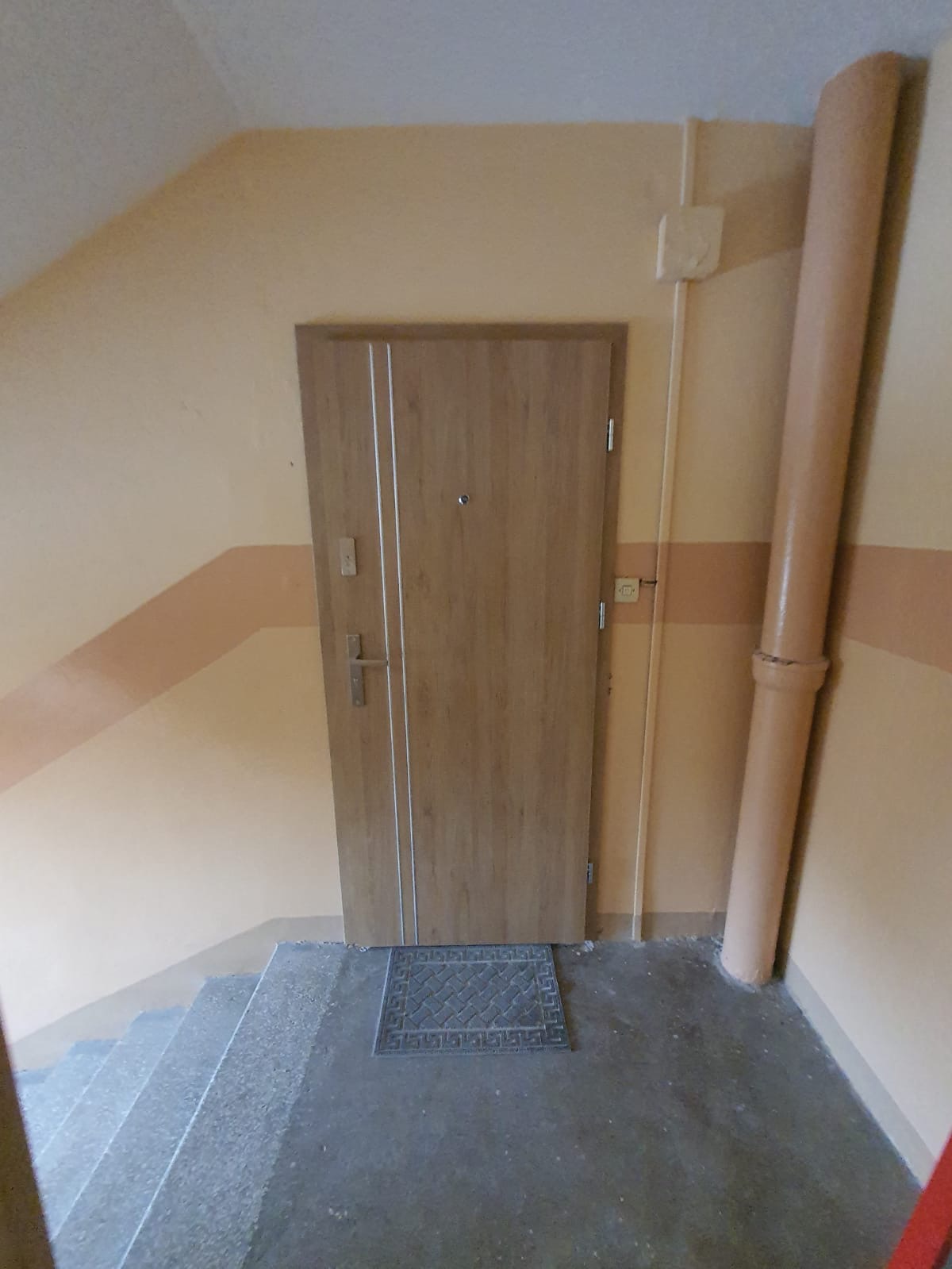 Zamontowane, drewnopodobne drzwi wejściowe z dwoma pionowymi, srebrnymi paskami, widoczne od strony klatki schodowej z szarymi schodami i beżowymi ścianami z brązowym pasem oraz rurą.
