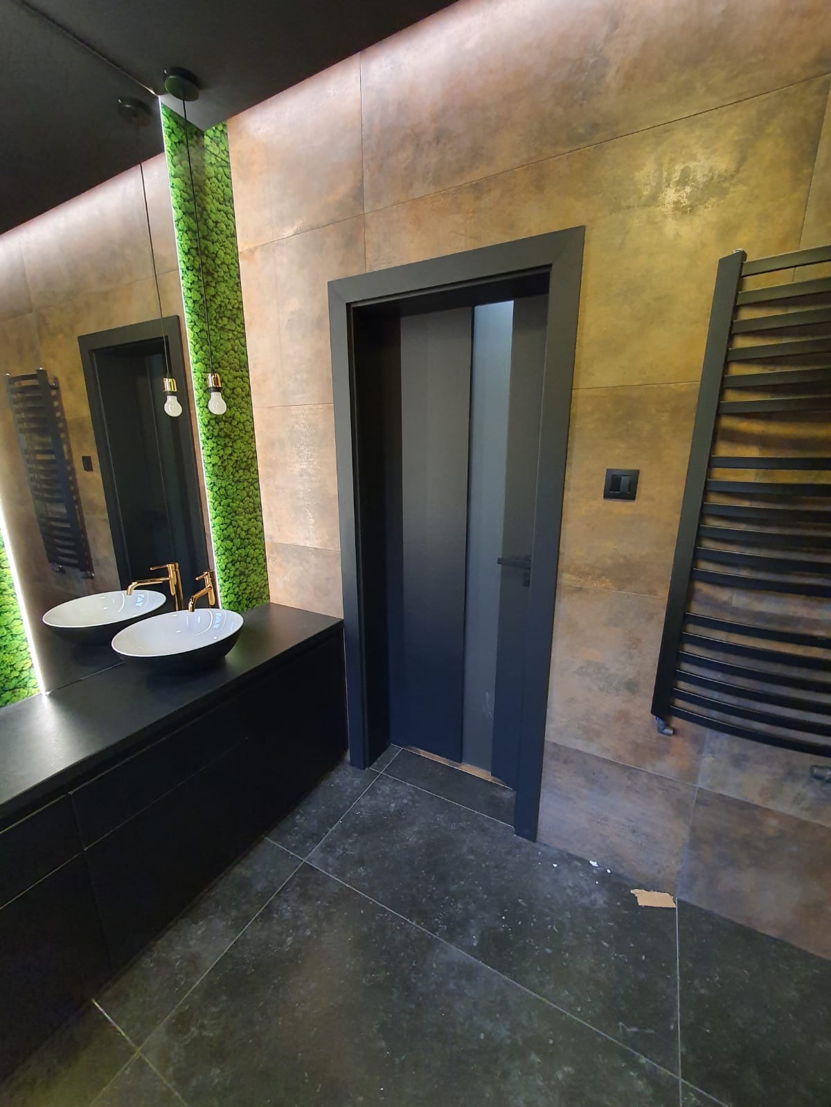 Nowoczesna łazienka z czarną umywalką nablatową, złotą armaturą, lustrem z roślinną dekoracją, ciemnymi płytkami podłogowymi i przesuwnymi drzwiami w czarnej ramie.