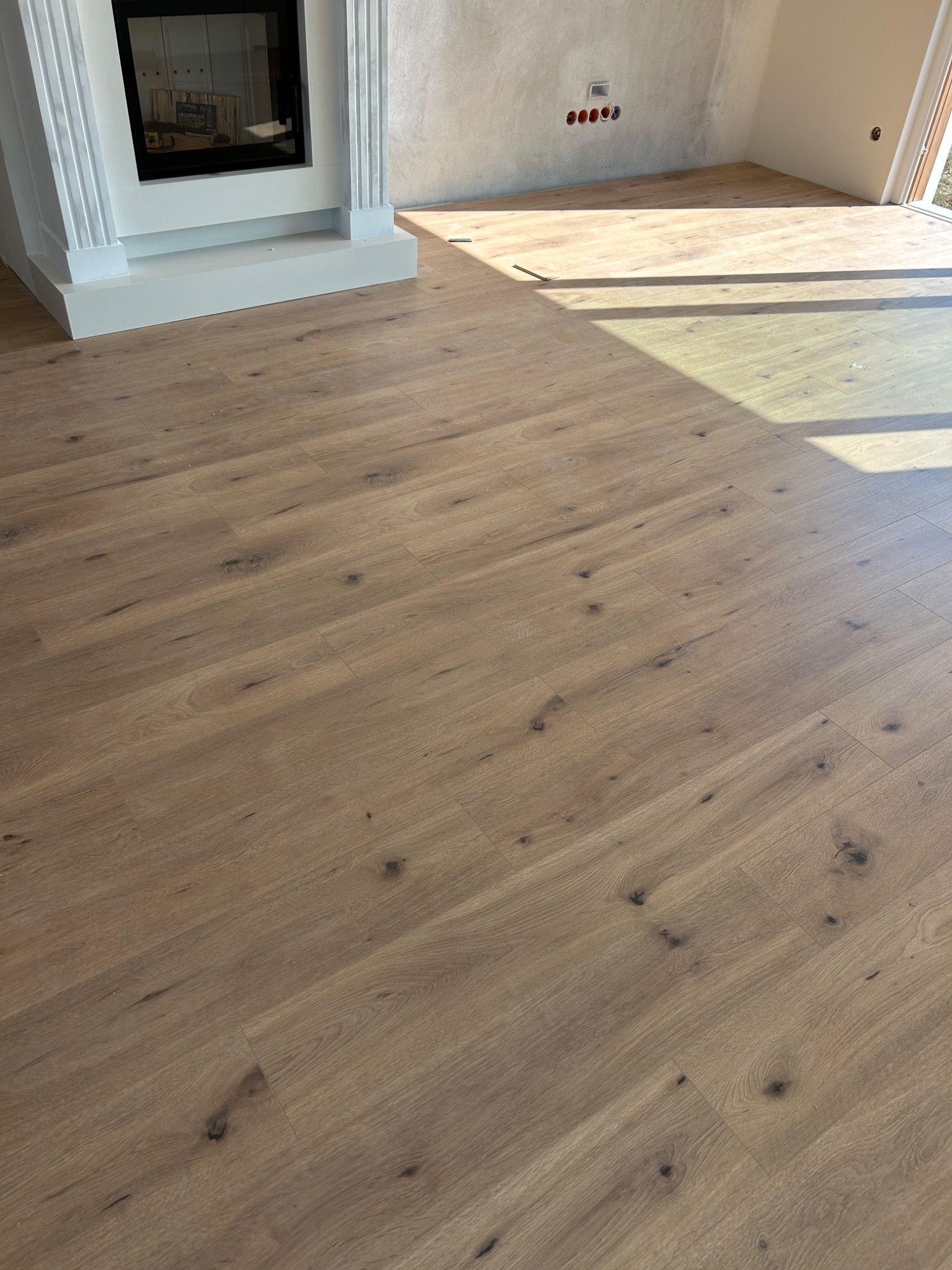 Jasne, drewniane deski podłogowe ułożone w nowym pomieszczeniu z kominkiem i surowymi ścianami, wpadające promienie słoneczne.