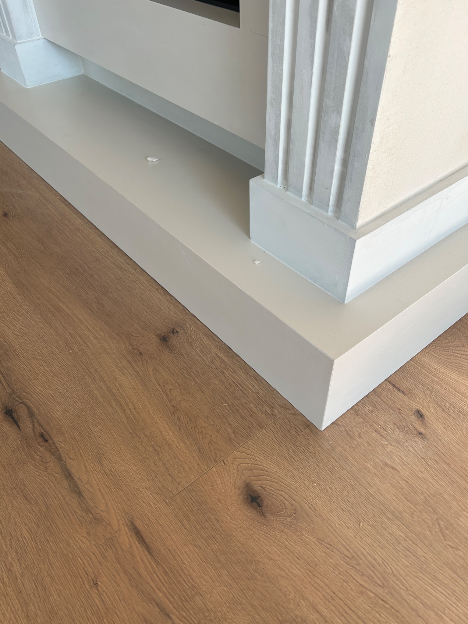 Fragment ułożonej podłogi z paneli drewnopodobnych przy białej obudowie kominka z dekoracyjnymi pilastrami, widoczne drobne zabrudzenia.