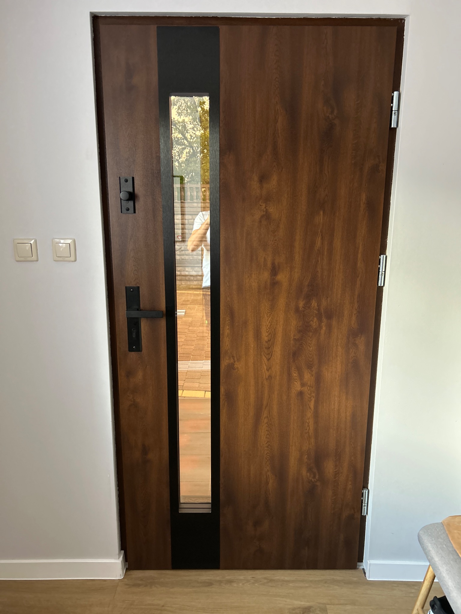 Brązowe drzwi wejściowe z czarnym, pionowym przeszkleniem, widoczne zamek i klamka, zamontowane w białej ścianie.