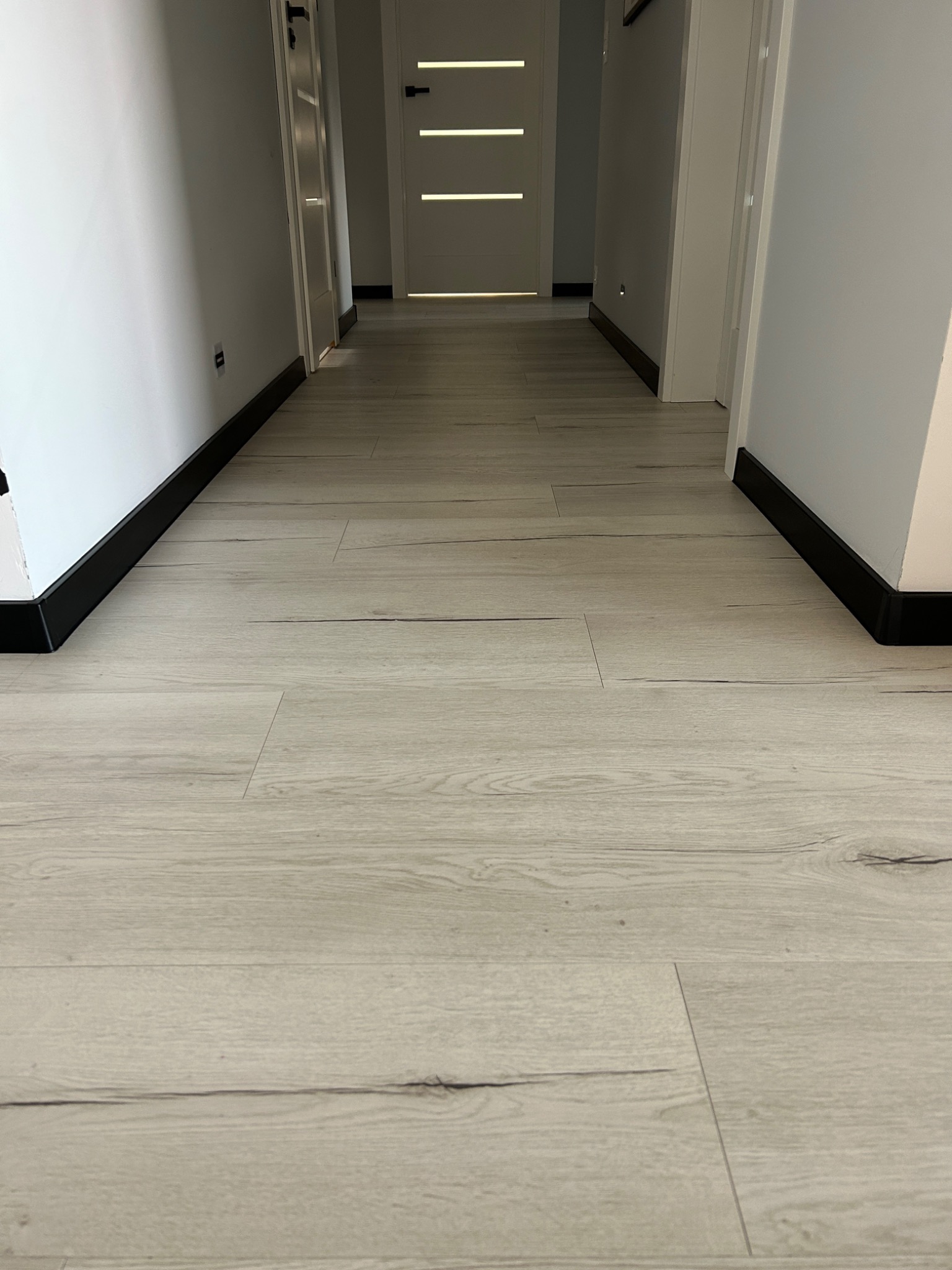 Jasne panele podłogowe ułożone w korytarzu z białymi ścianami i czarnymi listwami przypodłogowymi, widok w perspektywie.