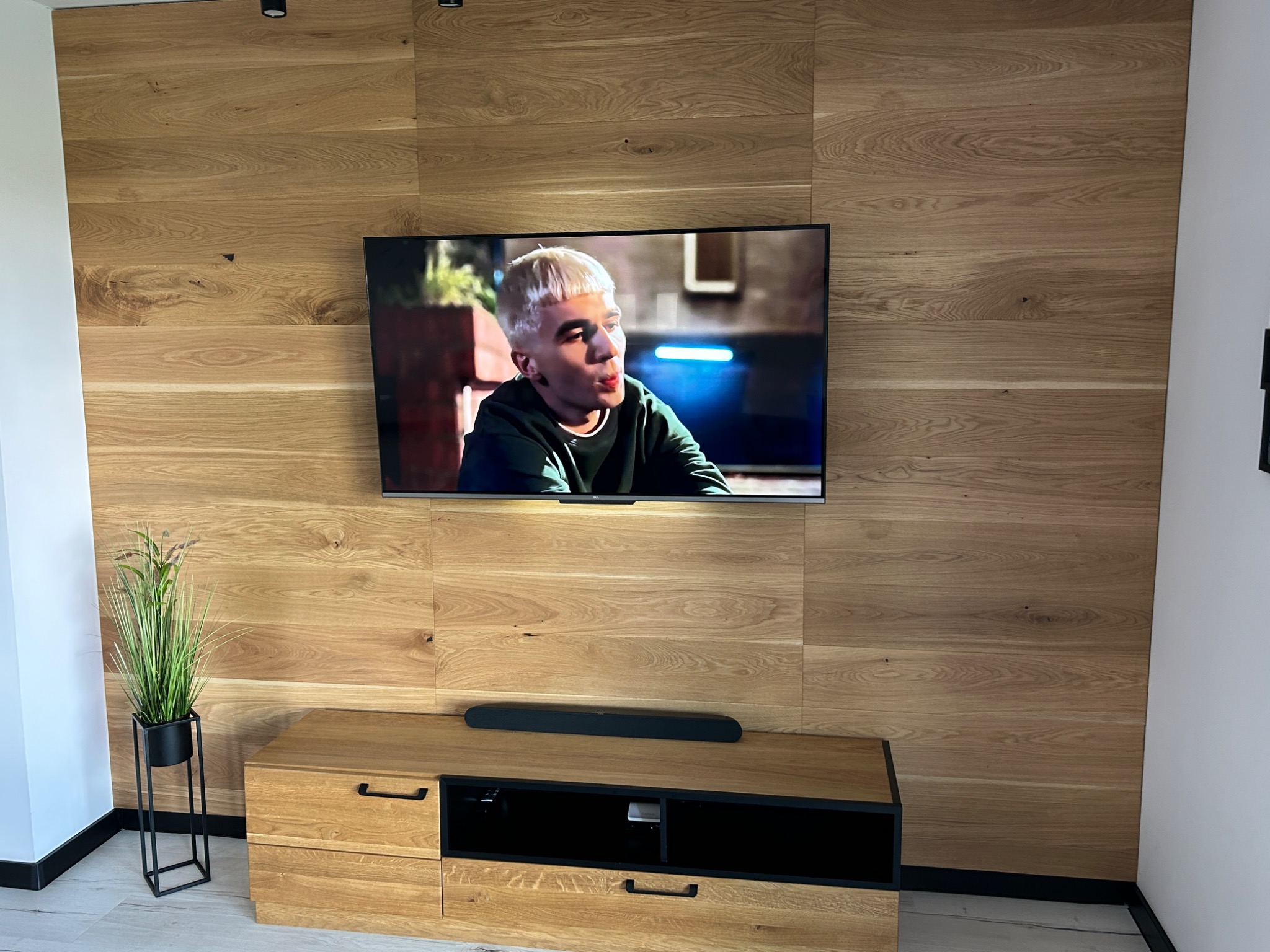 Drewniana ściana z poziomych paneli z telewizorem, szafką RTV i dekoracyjną trawą w czarnej doniczce.