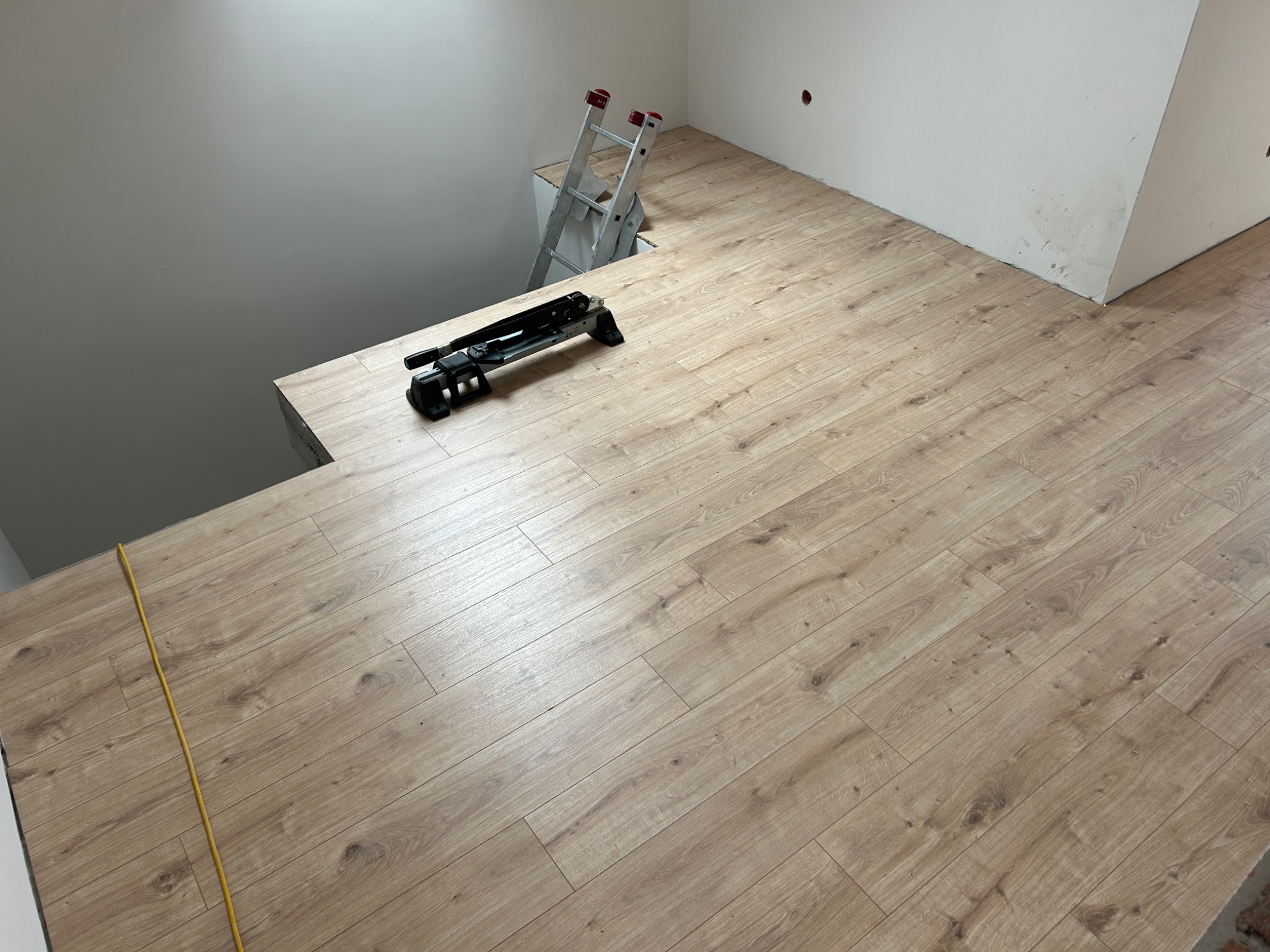 Fragment pomieszczenia z ułożonymi panelami podłogowymi w odcieniach beżu i brązu, widoczna drabina aluminiowa oparta o ścianę i gilotyna do cięcia paneli.