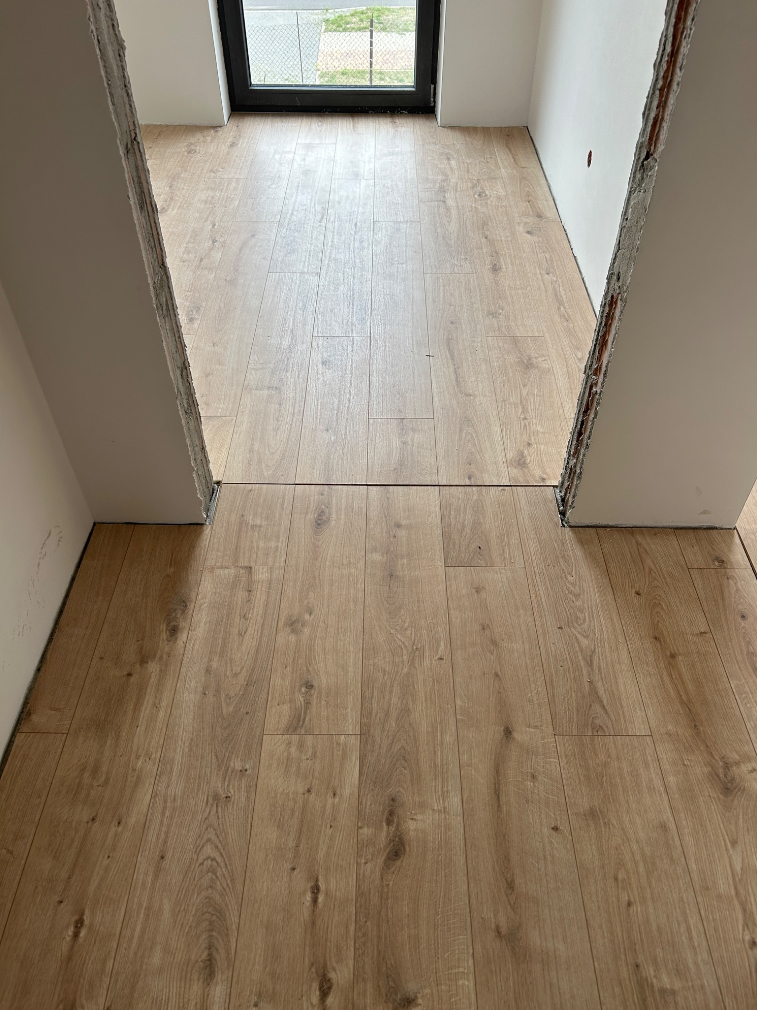 Jasne panele podłogowe ułożone w przejściu między pokojami, widoczne surowe krawędzie ścian przed wykończeniem.