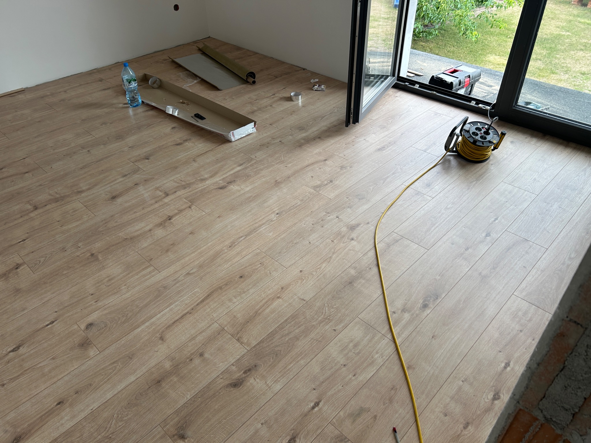 Jasne panele podłogowe ułożone w nowym pomieszczeniu z widocznymi materiałami montażowymi, butelką wody i zwiniętym przedłużaczem elektrycznym przy szklanych drzwiach balkonowych.