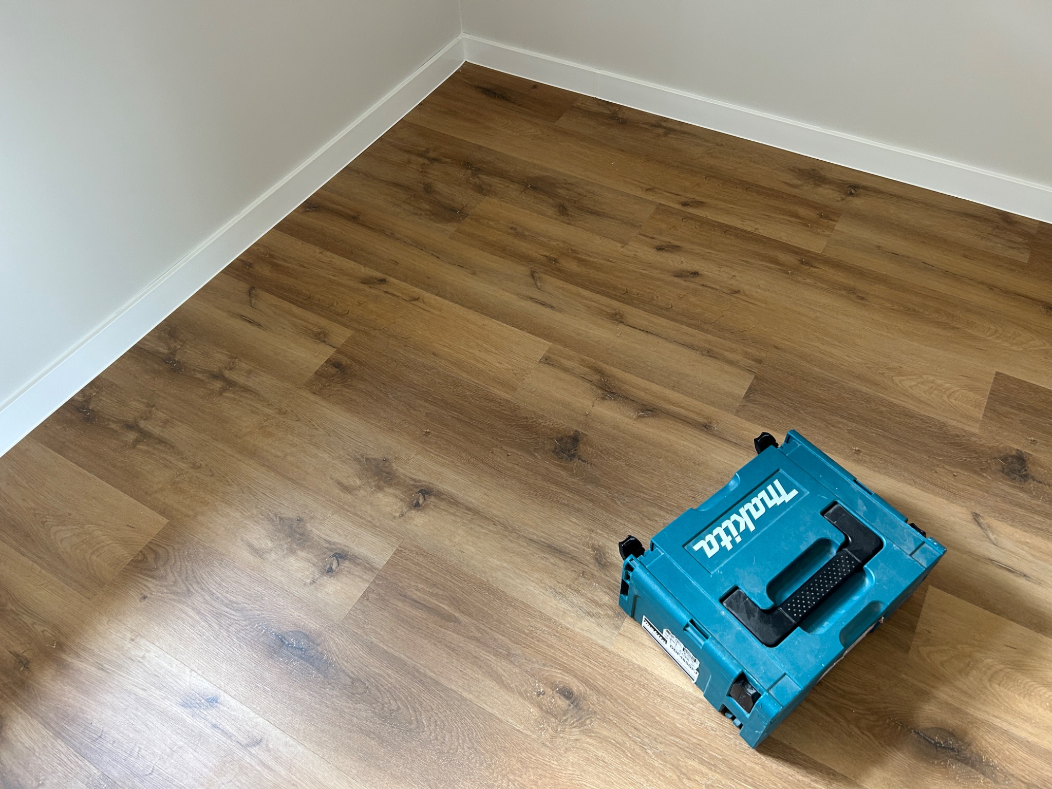 Świeżo ułożona podłoga z paneli laminowanych w odcieniach brązu, przy ścianie biała listwa przypodłogowa, obok niebieska skrzynka narzędziowa marki Makita.