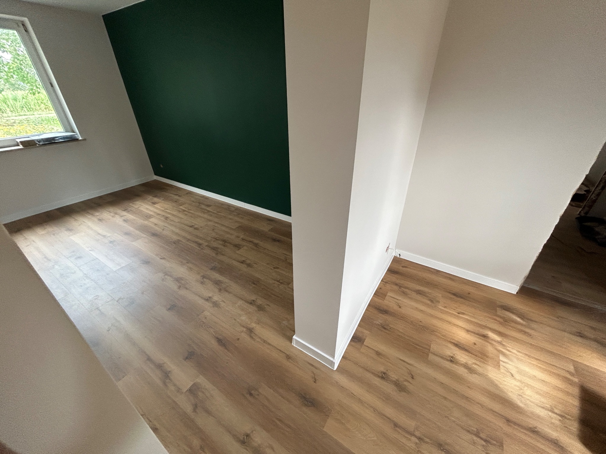 Jasne panele podłogowe w nowym pomieszczeniu z zieloną ścianą akcentującą, widoczne listwy przypodłogowe i okno z widokiem na zieleń.