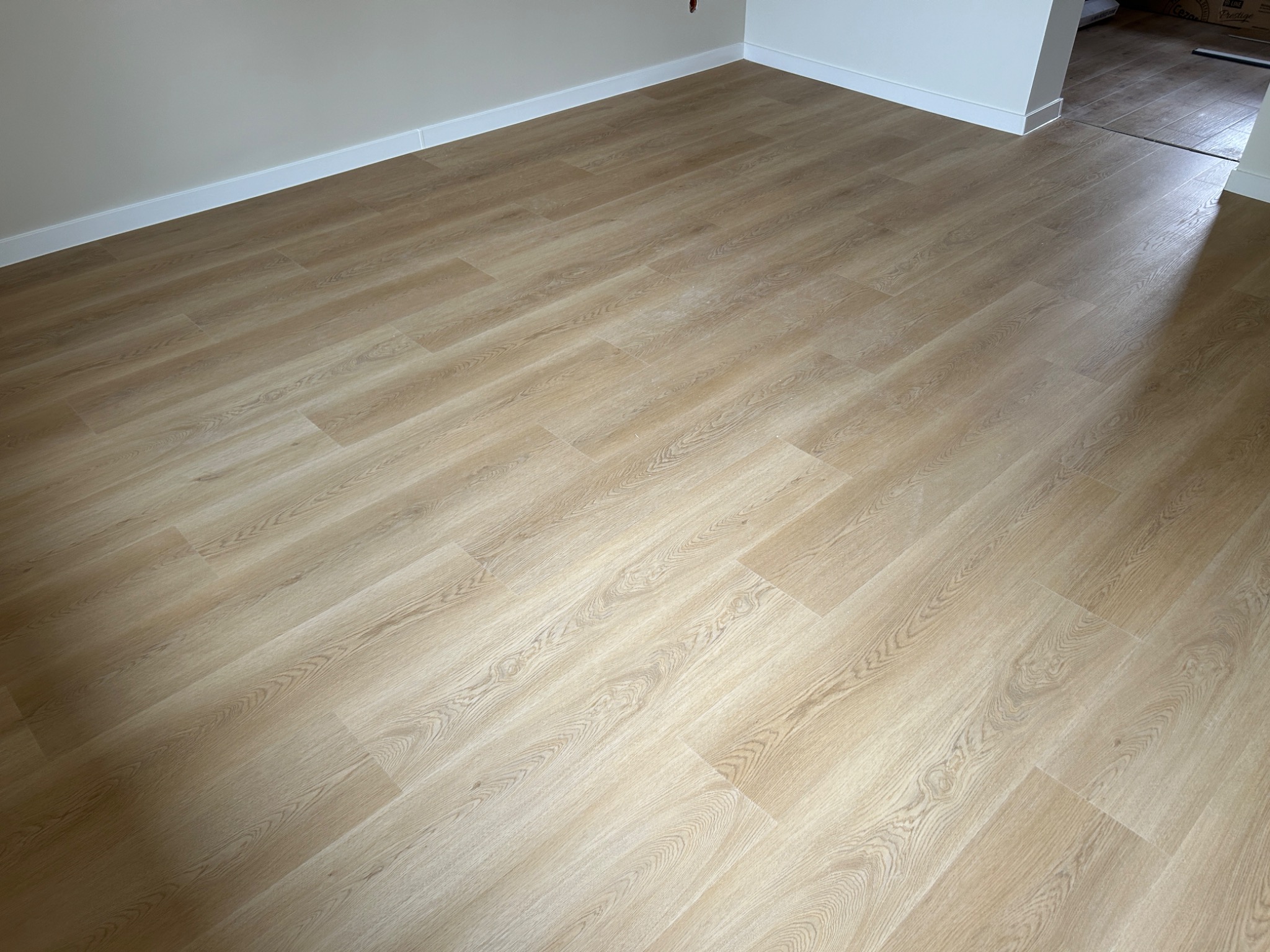 Jasne, drewnopodobne panele podłogowe ułożone w nowym pomieszczeniu, widoczne listwy przypodłogowe i fragment innego pomieszczenia w tle.