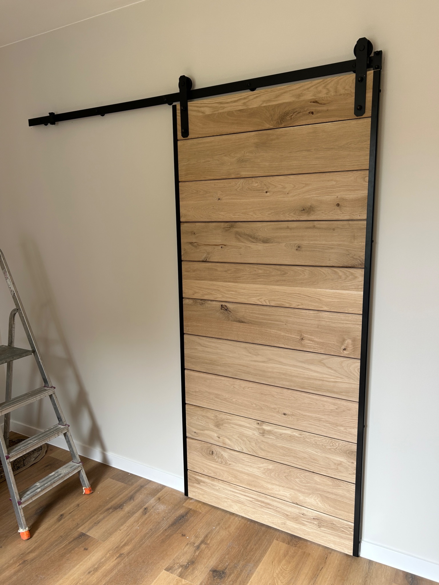 Zamontowane przesuwne drzwi drewniane z czarnymi okuciami na tle białej ściany, widoczna drabina malarska.