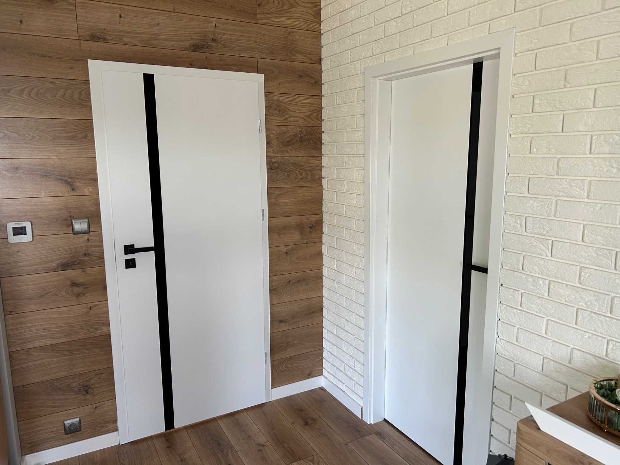 Dwa białe, nowoczesne drzwi wewnętrzne z czarnymi akcentami na tle ściany z paneli drewnopodobnych i ściany z białej cegły.