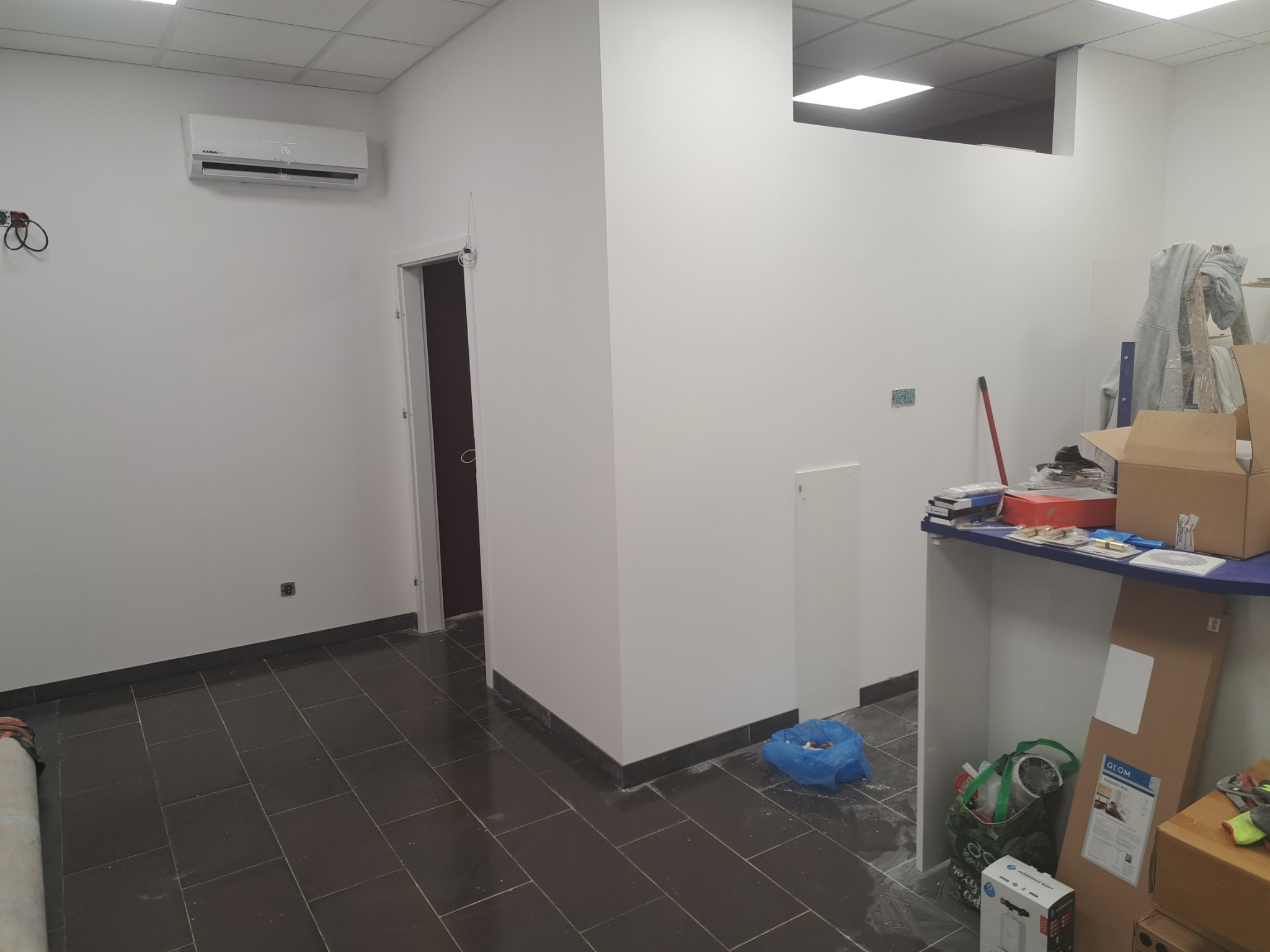 Wnętrze biura po remoncie, z pomalowanymi na biało ścianami, ciemną podłogą z płytek, klimatyzacją na ścianie i stertą materiałów budowlanych w rogu.
