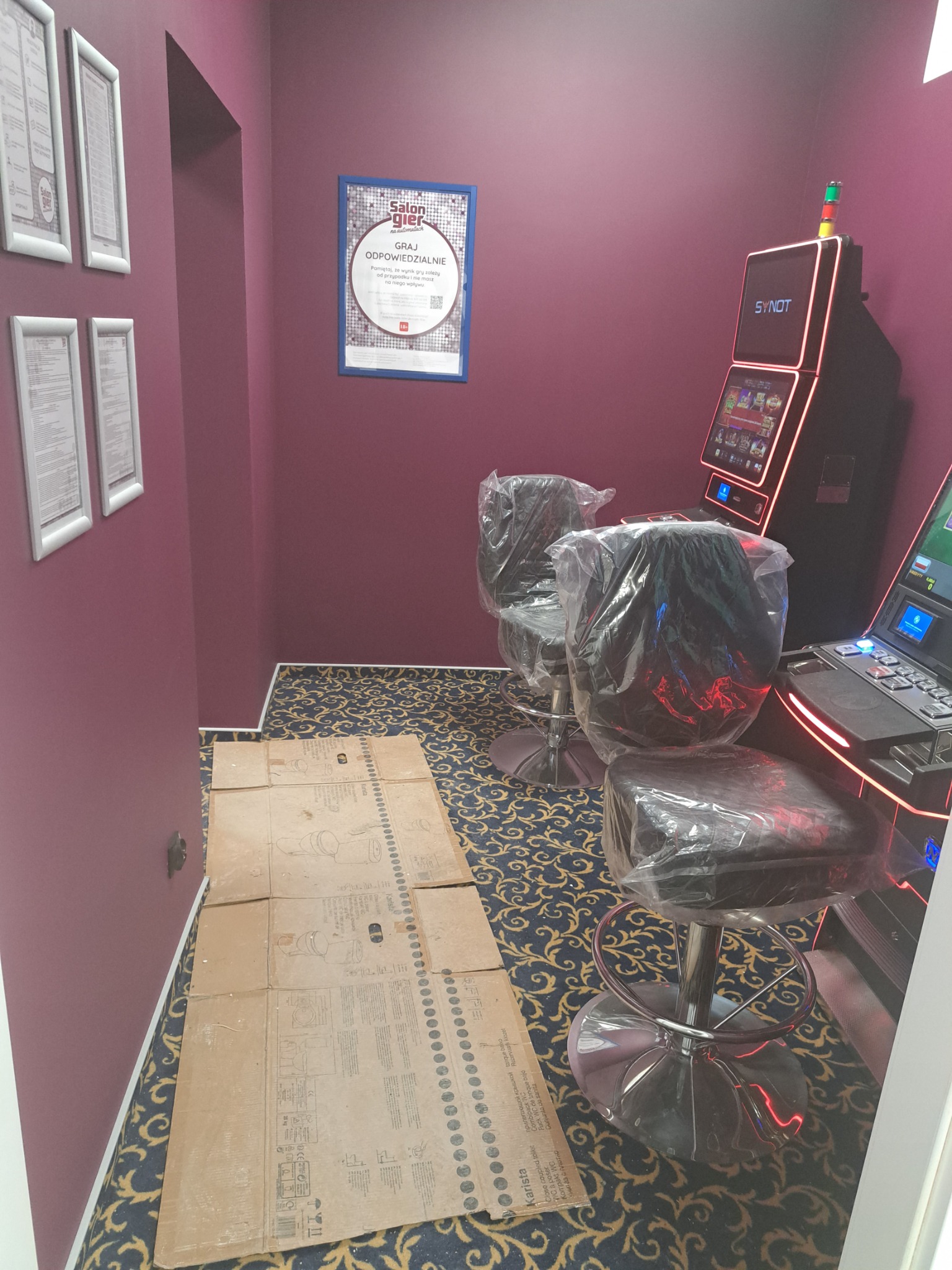 Przygotowanie do montażu automatów do gier: podłoga zabezpieczona kartonem, krzesła i automaty owinięte folią ochronną w lokalu z fioletowymi ścianami i wzorzystą wykładziną.