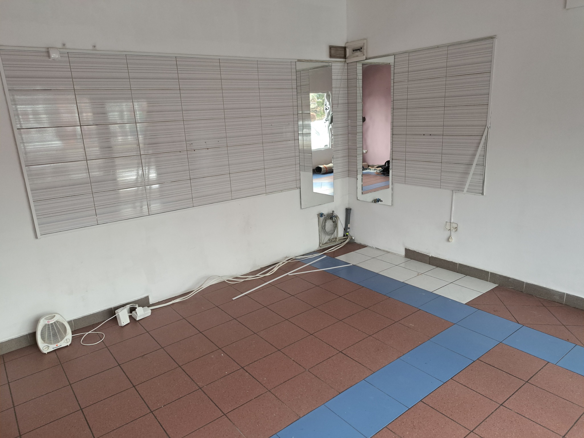 Wnętrze pomieszczenia w trakcie prac wykończeniowych: częściowo wyłożone płytkami ściany, podłoga z terakoty z niebieskim pasem, widoczne kable elektryczne, grzejnik elektryczny i dwa lustra.