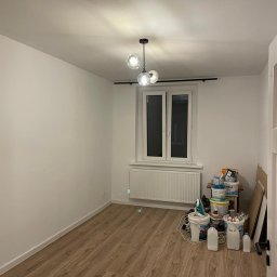 Dawid Wykończenia - Pusty pokój z drewnianą podłogą i białymi ścianami, przygotowany do prac wykończeniowych. Widoczne wiadra z farbą i inne materiały malarskie zgromadzone pod oknem.