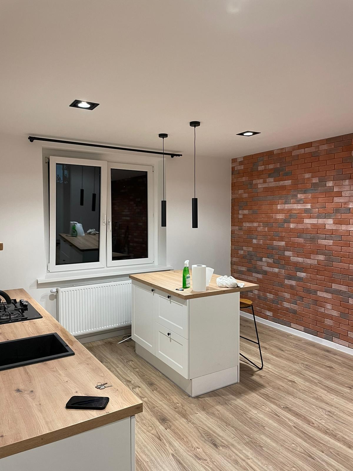 Nowoczesna kuchnia z wyspą, ceglaną ścianą i wiszącymi lampami cylindrycznymi; widoczny smartfon na blacie.