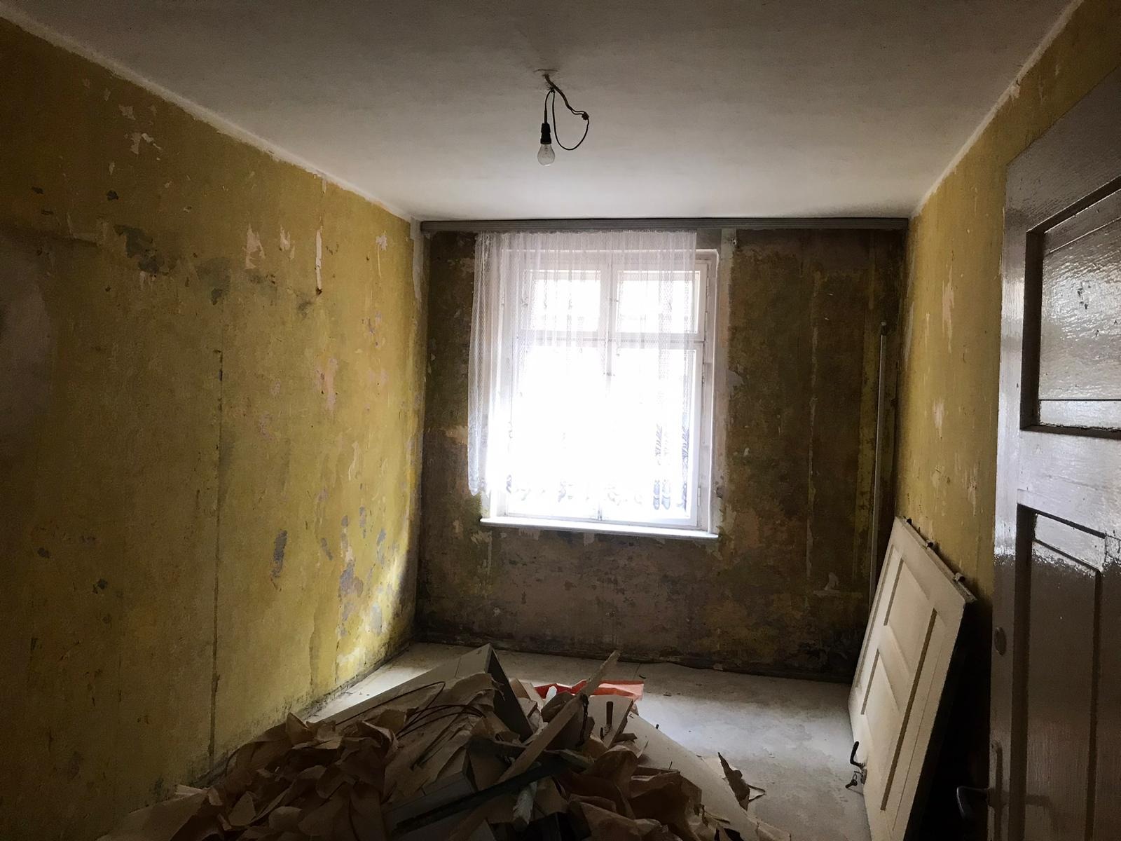 Pokój w trakcie remontu: ściany z resztkami starej farby, okno z firanką, żarówka na kablu zwisająca z sufitu, na podłodze sterta odpadów i oparte o ścianę drzwi.